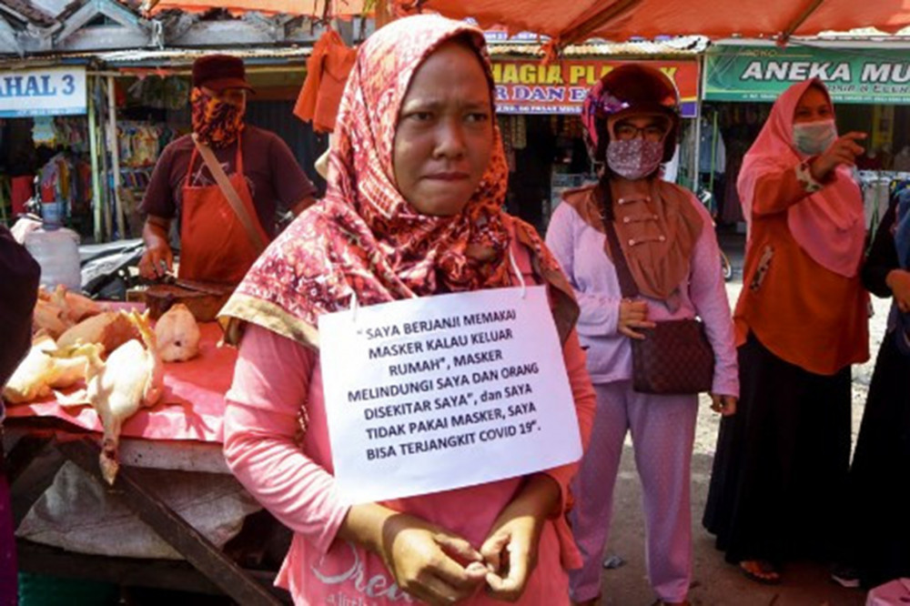 Tỉnh Bengkulu, 13/5/2020: Bức hình này, chụp một phụ nữ vi phạm bị buộc đứng ngoài phố với tấm bảng “Tôi hứa sẽ đeo khẩu trang” treo trước ngực, được tải lên phương tiện truyền thông xã hội để làm “bài học” giữa đại dịch Covid-19 ở Indonesia (AFP). Ở thủ đô Jakarta, những cư dân vi phạm các quy tắc giãn cách xã hội buộc phải mặc áo khoác “Người phá vỡ quy tắc” khi làm sạch các cơ sở công cộng, gồm cả các nhà vệ sinh. Ở tỉnh Aceh bảo thủ, những người vi phạm phải đọc các đoạn kinh Koran để chuộc lỗi. Người không theo đạo Hồi được miễn trừ hình phạt này.