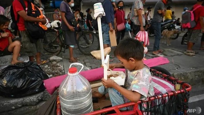 Một đứa trẻ vui mừng khi nhận phần cơm từ thiện, phía sau là dòng người vẫn đang xếp hàng chờ lấy thức ăn.