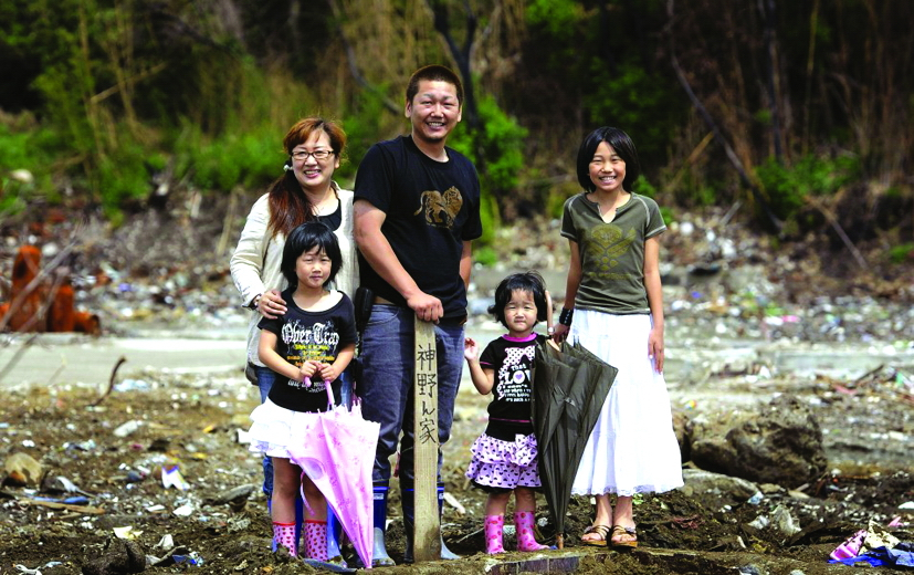 Fumitoshi Kamino và gia đình đứng tại vị trí từng là ngôi nhà cũ của họ ở Ishinomaki, tỉnh Miyagi, vào ngày 25/6/2011. Mười năm sau, những cô con gái của gia đình đã trưởng thành, dù trong thâm tâm, tất cả đều bị ám ảnh về ký ức thảm họa -  Ảnh: Kyodo