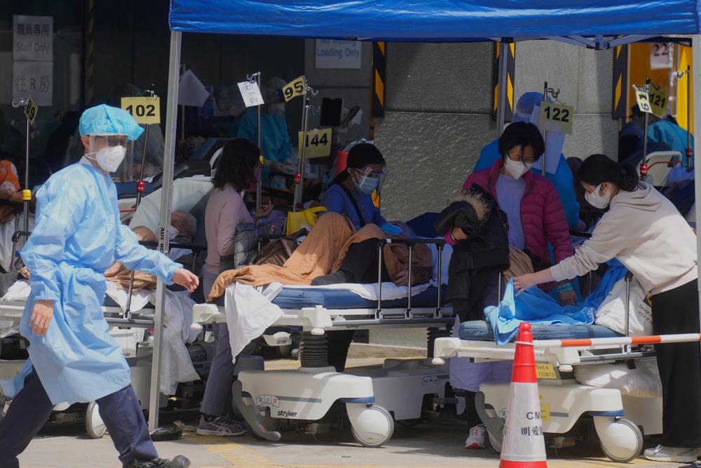 Bệnh nhân trên giường bệnh chờ đợi tại khu vực tạm giữ bên ngoài Trung tâm Y tế Caritas ở Hồng Kông vào ngày 28 tháng 2 năm 2022. Các biến thể omicron lây lan nhanh chóng đang áp đảo Hồng Kông, khiến cho việc kiểm tra hàng loạt, kiểm dịch, mua hàng trong siêu thị và sự thiếu hụt giường bệnh. Ngay cả những nhà xác cũng tràn ngập, buộc các cơ quan chức năng phải cất giữ các thi thể trong các container vận chuyển lạnh. (Ảnh AP / Vincent Yu, Tệp)