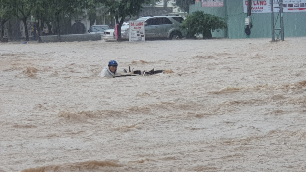 1 người đàn ông dắt xe trên đường Hàn Mặc Tử (phường Ghềnh Ráng) bị nước lũ cuốn, người dân phải dăng dây để hỗ trợ người này vào khu vực an toàn.