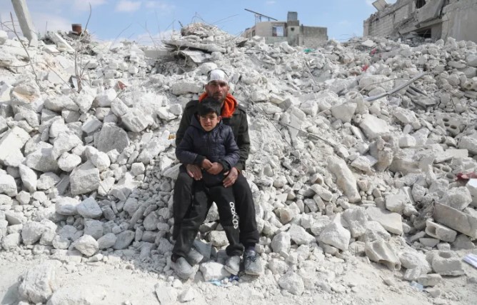 Mahmoud Omar al-Ormi ngồi cùng con trai Ahmad trên đống đổ nát của tòa nhà ở Atarib, tây bắc Syria. Al-Ormi đã mất người vợ đang mang thai và bốn đứa con vì trận động đất [Ali Haj Suleiman/Al Jazeera]
