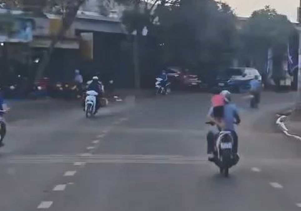 Người đàn ông một tay đi xe máy, một tay giữ bé gái đứng nghiêng ngã sau yên xe