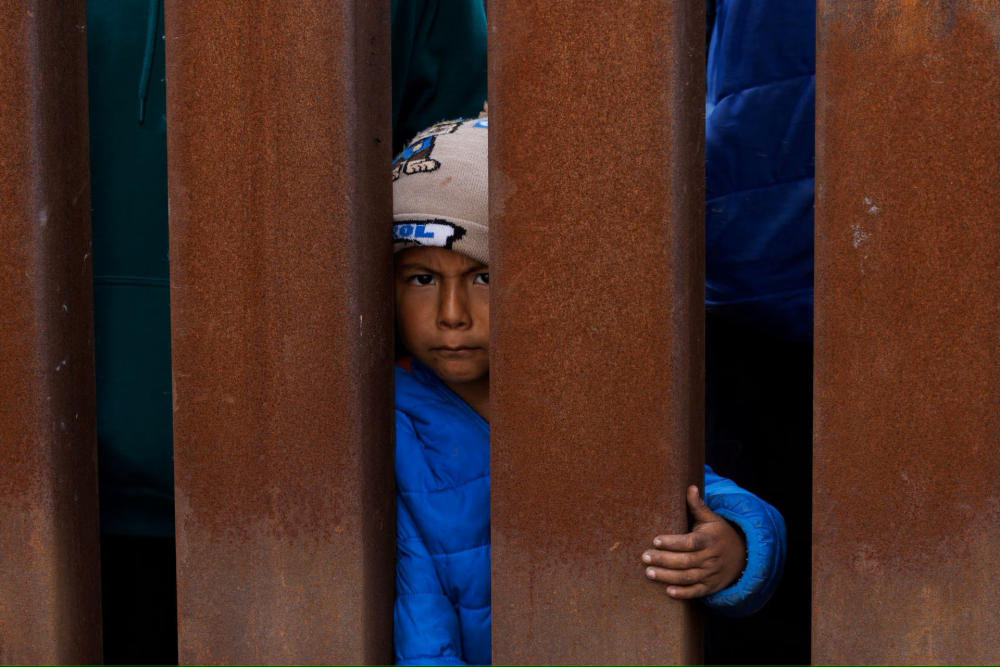 Một cậu bé di cư chờ thức ăn ở hàng rào biên giới trong khi những người di cư tụ tập giữa hàng rào biên giới chính và phụ, gần San Diego, California. (Ảnh: REUTERS)