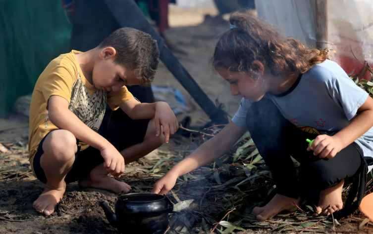 Những đứa trẻ Palestine di tản trong nước, những người chạy trốn khỏi cuộc bắn phá của Israel ở phía bắc Gaza và hiện đang sống trong những nơi trú ẩn tạm bợ, đốt lửa để đun ấm nước sau cơn mưa ở Khan Younis vào ngày 15 tháng 11 năm 2023. [Mahmud Hams/AFP]