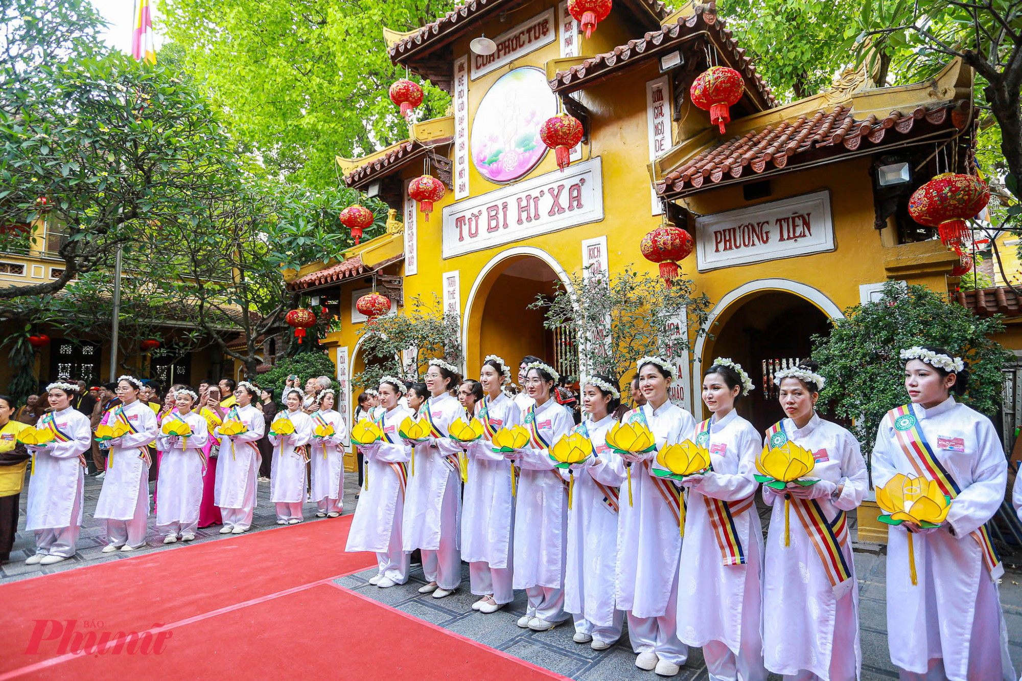 Phật tử trong trang phục chỉnh tề, tay cầm cờ và hoa sen, thành kính xếp hàng trước cổng chùa.