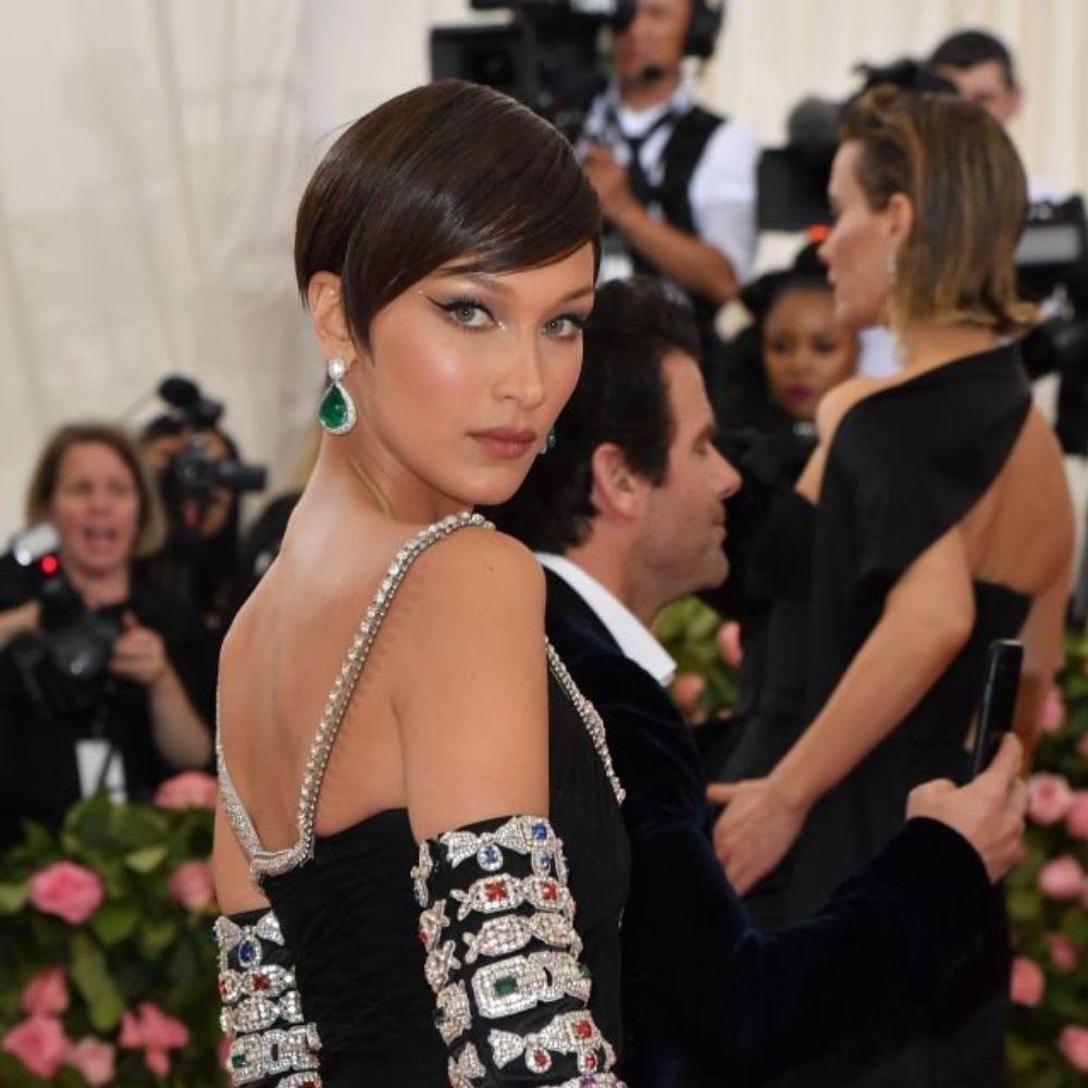 Bella Hadid đã thu hút mọi ánh nhìn với mái tóc pixie cổ điển, ôm sát đầu (sleek vintage pixie) đầy quyến rũ. Mái tóc được tạo kiểu mượt mà, ép bằng lược tròn và máy sấy, phần mái được vuốt lệch sang một bên kết hợp cùng tóc mai nhọn và những đường cắt sắc nét. Tổng thể mang đến một vẻ ngoài retro sang trọng, gợi nhớ đến phong cách Hollywood những năm 1950.  Kiểu tóc pixie này đặc biệt lý tưởng cho những ai sở hữu gương mặt dài và góc cạnh, vì phom tóc ôm sát sẽ làm nổi bật những đường nét khuôn mặt, tạo cảm giác thanh thoát và sắc sảo. Phần mái vắt chéo giúp cân bằng tỷ lệ khuôn mặt, làm mềm mại và giảm bớt sự góc cạnh của vùng trán cao hoặc rộng.