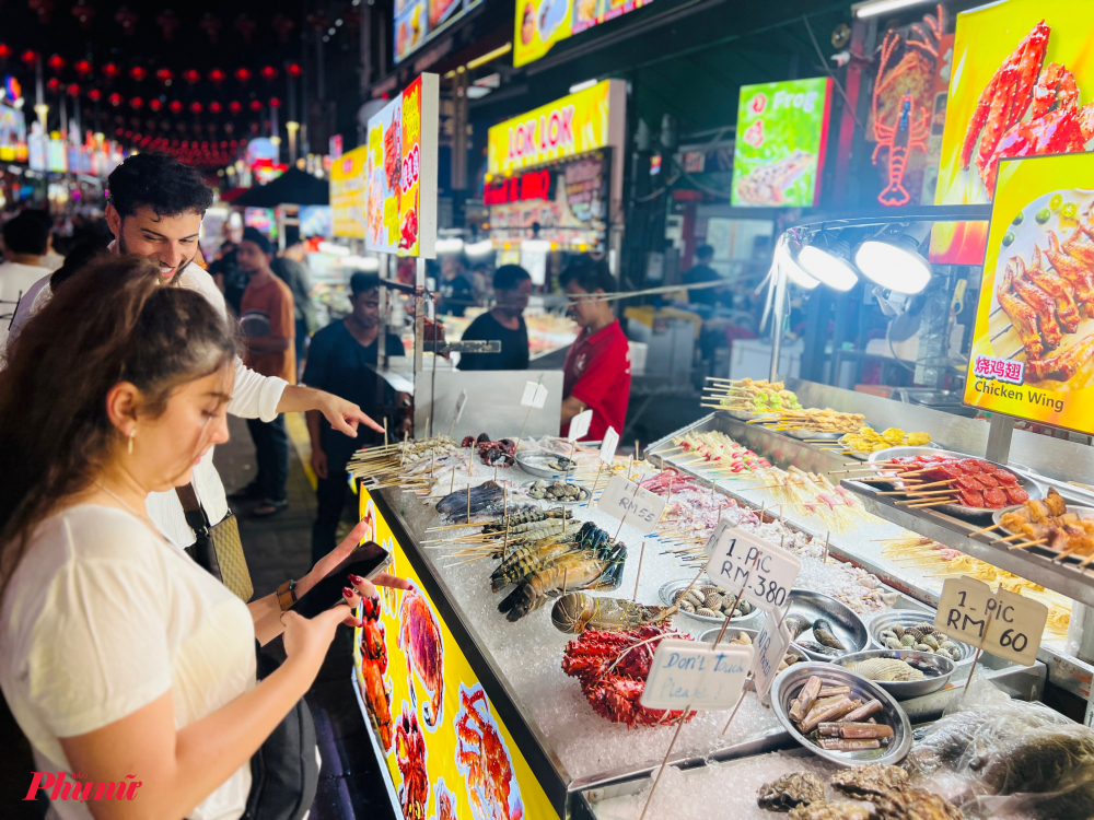 Chỉ mất chưa đầy 2 giờ bay từ TP.HCM, du khách Việt đã có thể đặt chân đến Kuala Lumpur – thủ đô sôi động của Malaysia, nơi những khu Pasar Malam (chợ đêm) rực rỡ ánh đèn là điểm hẹn không thể bỏ lỡ để khám phá đời sống và văn hóa bản địa.  Giữa vô vàn chợ đêm sôi động, Jalan Alor Food Street là cái tên nổi bật, được ví như thiên đường ẩm thực đêm giữa trung tâm Bukit Bintang. Từ 14h30 chiều đến tận 3h sáng hôm sau, con phố này bừng sáng bởi hàng trăm gian hàng bán đủ loại đặc sản Malaysia như cơm Nasi Lemak, cơm chiên Nasi Goreng, thịt xiên Satay, trà sữa kéo Teh Tarik,... Đặc biệt, sầu riêng Musang King – niềm tự hào quốc gia – là món nhất định phải thử. Ngoài ra, thực khách còn có thể thưởng thức các món ăn mang hương vị Việt Nam, Trung Quốc, Thái Lan, Ấn Độ,... với giá dao động chỉ từ 10–15 RM/món (khoảng 55.000 – 70.000 đồng).  Pasar Malam không chỉ là nơi ăn uống, mà còn là nơi thể hiện rõ nét nhất sự giao thoa văn hóa của quốc gia đa sắc tộc Malaysia. Không khí tấp nập, mùi hương ẩm thực quyện trong tiếng cười nói rộn ràng chính là lý do khiến những khu chợ đêm như Jalan Alor trở thành linh hồn của Kuala Lumpur.  Nắm bắt sức hút này, ngành du lịch Malaysia đang đẩy mạnh kết nối với Việt Nam, khuyến khích mở thêm các đường bay thẳng. Chỉ riêng trong những tháng đầu năm 2025, Malaysia đã đón hơn 54.000 lượt khách Việt – con số ấn tượng phản ánh nhu cầu khám phá ngày càng cao.  Hiện nay, Vietnam Airlines đang khai thác đều đặn 14 chuyến/tuần giữa TP.HCM – Kuala Lumpur và Hà Nội – Kuala Lumpur bằng các dòng máy bay hiện đại như Airbus A321/A321NEO. Hãng cũng cam kết tiếp tục mở rộng mạng bay và nâng cao dịch vụ, góp phần kết nối hiệu quả giữa Việt Nam và Malaysia – điểm đến chiến lược tại Đông Nam Á.