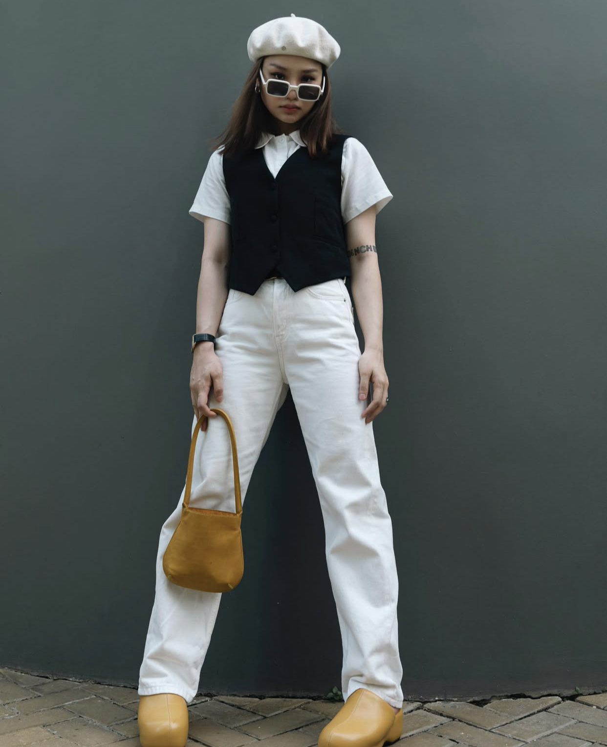 gu thời trang phóng khoáng pha chút cổ điển. Cô phối sơ mi trắng và áo gile đen cùng quần jeans trắng, tạo nên tổng thể tối giản nhưng nổi bật. Mũ beret, túi xách và giày vàng mù tạt là điểm nhấn thú vị, mang lại sắc màu trẻ trung, cá tính và đậm chất nghệ sĩ đường phố.