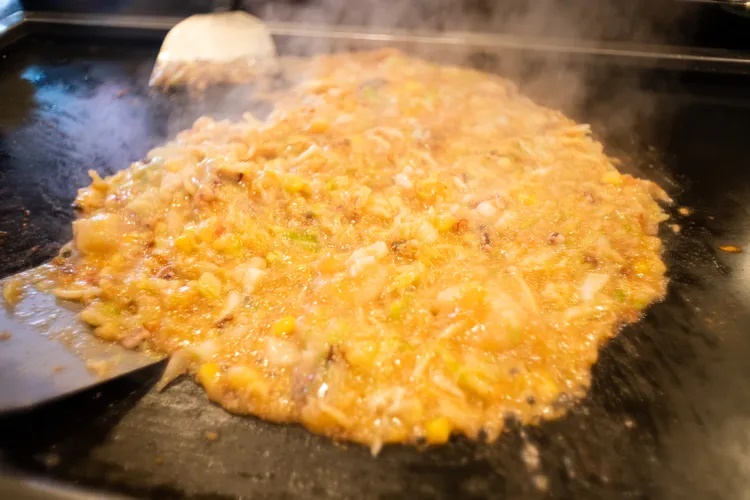 Ăn monjayaki ở Tsukishima. Monjayaki là một loại bánh xèo áp chảo kiểu Nhật. Khi áp chảo, vỏ bánh phía dưới giòn, phần trên mềm, ẩm và hơi chảy. Khách thường tự chế biến bánh trên chảo nóng đặt tại bàn. Monjayaki có hương vị của sốt, rau củ và các loại nhân. Trên đảo nhân tạo Tsukishima, hàng chục nhà hàng monjayaki tập trung ở phố Nishi-Nakadori. Ảnh trên: Monjayaki đang được áp chảo - Ảnh: Kayoko Hayashi/Getty Images