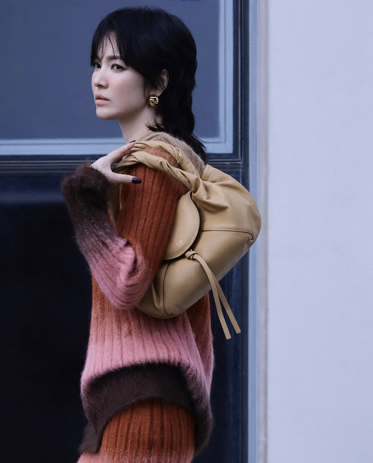 Song Hye Kyo Trang phục len dệt ombre phối màu hồng, nâu và đỏ đất tạo nên tổng thể ấm áp nhưng vẫn thời thượng, kết hợp cùng chiếc túi đeo vai màu be và khuyên tai bản tròn mạ vàng, tôn lên vẻ sang trọng, tinh tế.  Dù trong tạo hình phá cách, Song Hye Kyo vẫn giữ nguyên khí chất cuốn hút