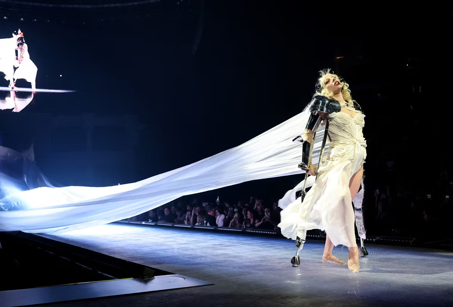 Mugler: Phần hai của concert mở ra với hình ảnh Gaga trong váy trắng phát sáng, tóc bạch kim, đi chân trần và chống nạng chrome – biểu tượng từ MV Paparazzi năm 2008. Nếu khi xưa cây nạng đại diện cho sự hy sinh vì danh tiếng, thì giờ đây, nó trở thành biểu tượng của sinh tồn. Gaga khập khiễng tiến về phía trước như một nữ chiến binh từng gục ngã nhưng không buông xuôi. Hình ảnh này là minh chứng sống động cho quá trình vượt qua bệnh tật thể chất lẫn tinh thần suốt hơn một thập kỷ của nữ ca sĩ.