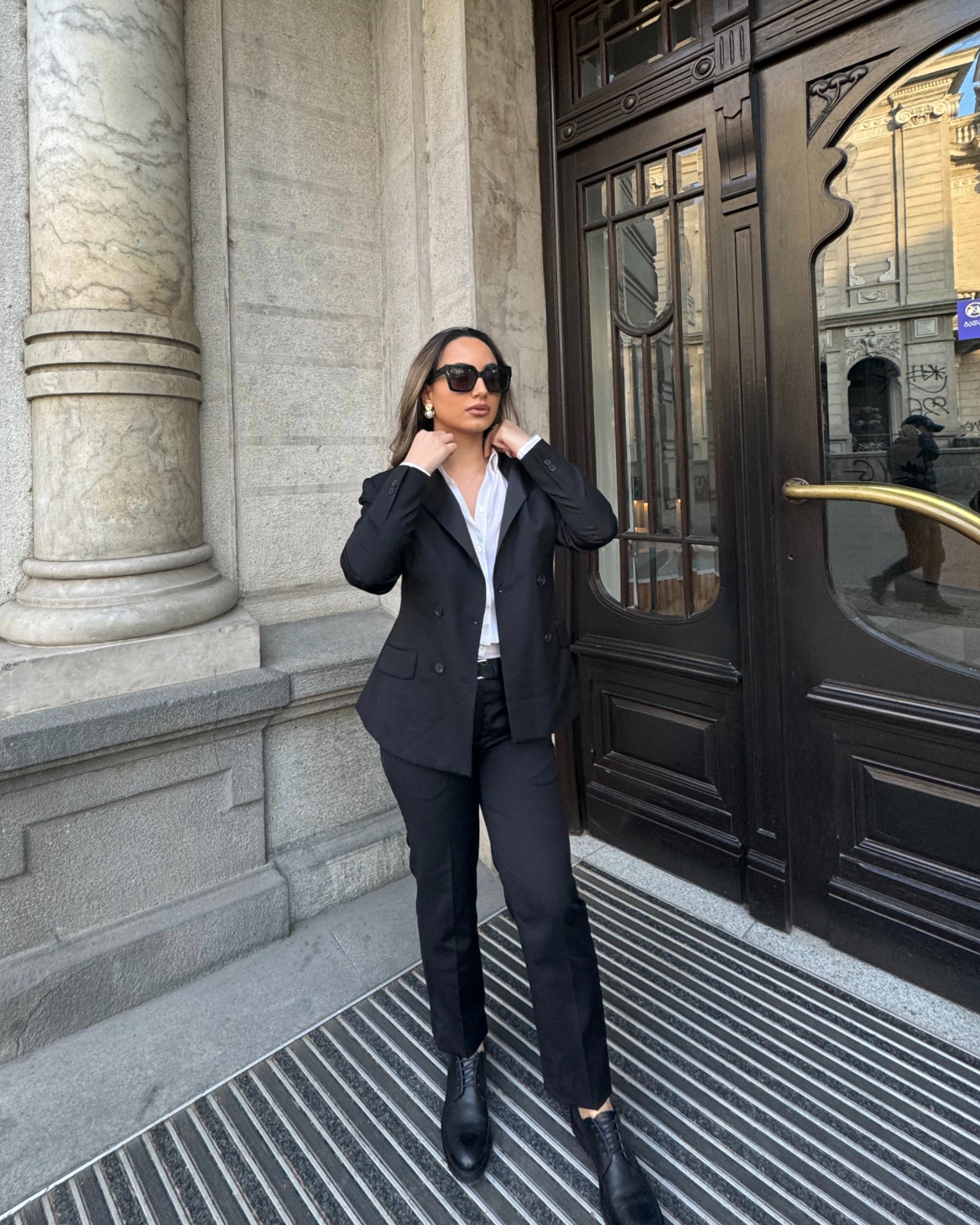 Bộ suit đen hai hàng khuy kết hợp sơ mi trắng mang đến diện mạo thanh lịch, chuyên nghiệp nhưng không kém phần cuốn hút. Đôi giày da đen cổ điển cùng kính râm to bản tạo điểm nhấn sang trọng, giúp tổng thể outfit thêm phần sắc sảo và hiện đại. 