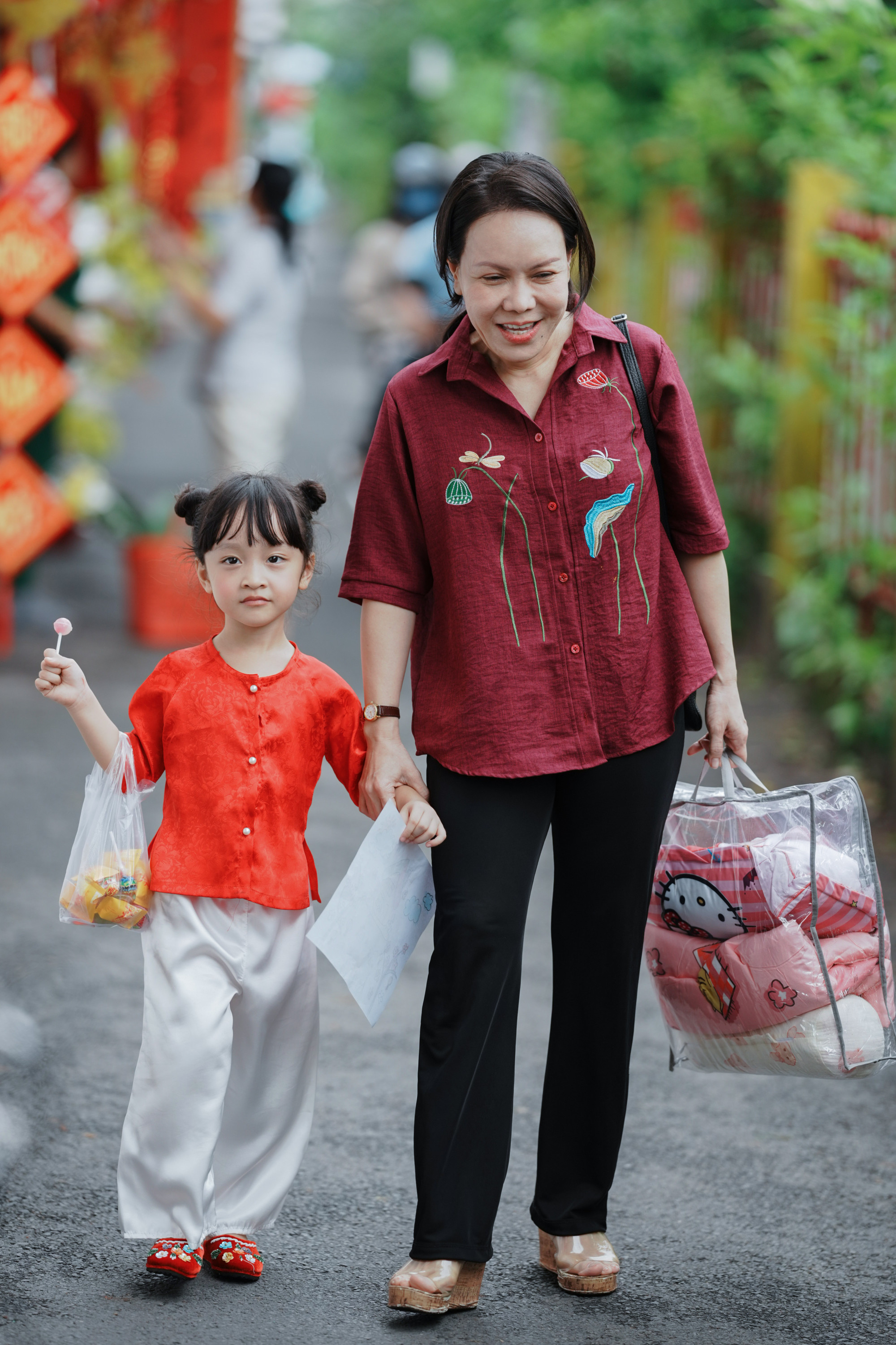 Trong phim, Việt Hương vào vai bà Hậu, thương con cháu hết mực nhưng đôi khi cứng nhắc. Nữ diễn viên thường xuất hiện với phong cách giản dị, gần gũi, phù hợp với hình ảnh một người mẹ, người bà tảo tần. Cô chọn áo sơ mi đỏ thẫm chất liệu thô mộc, điểm xuyết họa tiết thêu tay tinh tế như hoa sen và lá, tạo cảm giác mộc mạc nhưng vẫn duyên dáng. Quần đen ống suông kết hợp đôi guốc mộc càng tôn lên nét thân thuộc.