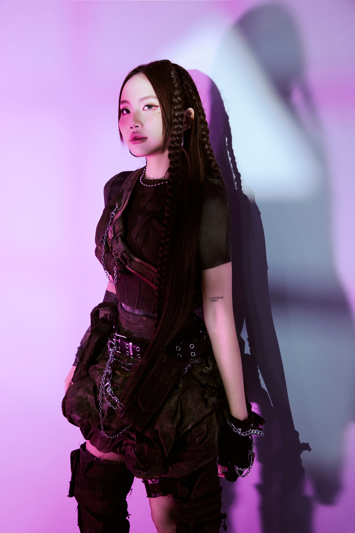 Phương Mỹ Chi xuất hiện với phong cách chiến binh hiện đại: outfit tông tối, corset ôm dáng, nhiều lớp xếp bất đối xứng, dây xích kim loại tạo cảm giác mạnh mẽ. Tóc tết dài, khuyên tai bản lớn cùng ánh sáng tím–hồng mang vibe cyberpunk, thể hiện hình ảnh nổi loạn, phá cách khác hẳn sự ngọt ngào trước đây.