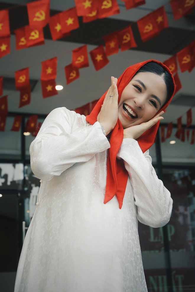 Hoa hậu Ngọc Hân mang đến một phong cách tươi trẻ, giản dị nhưng đầy tính truyền thống và đậm đà bản sắc Việt. Cô diện áo dài trắng cổ điển với chất liệu gấm nhẹ, vừa thanh lịch vừa thuần khiết. Chiếc khăn quàng đỏ được đội lên đầu theo phong cách thiếu nhi xưa tạo nên điểm nhấn thú vị, gợi nhớ hình ảnh học sinh trong những ngày lễ lớn của đất nước.  Nụ cười rạng rỡ và biểu cảm nhí nhảnh khiến tổng thể phong cách của Ngọc Hân vừa gần gũi, thân thiện, vừa thể hiện niềm tự hào và tinh thần lạc quan. Phông nền với những lá cờ đỏ sao vàng càng làm nổi bật tinh thần dân tộc trong trang phục của cô.
