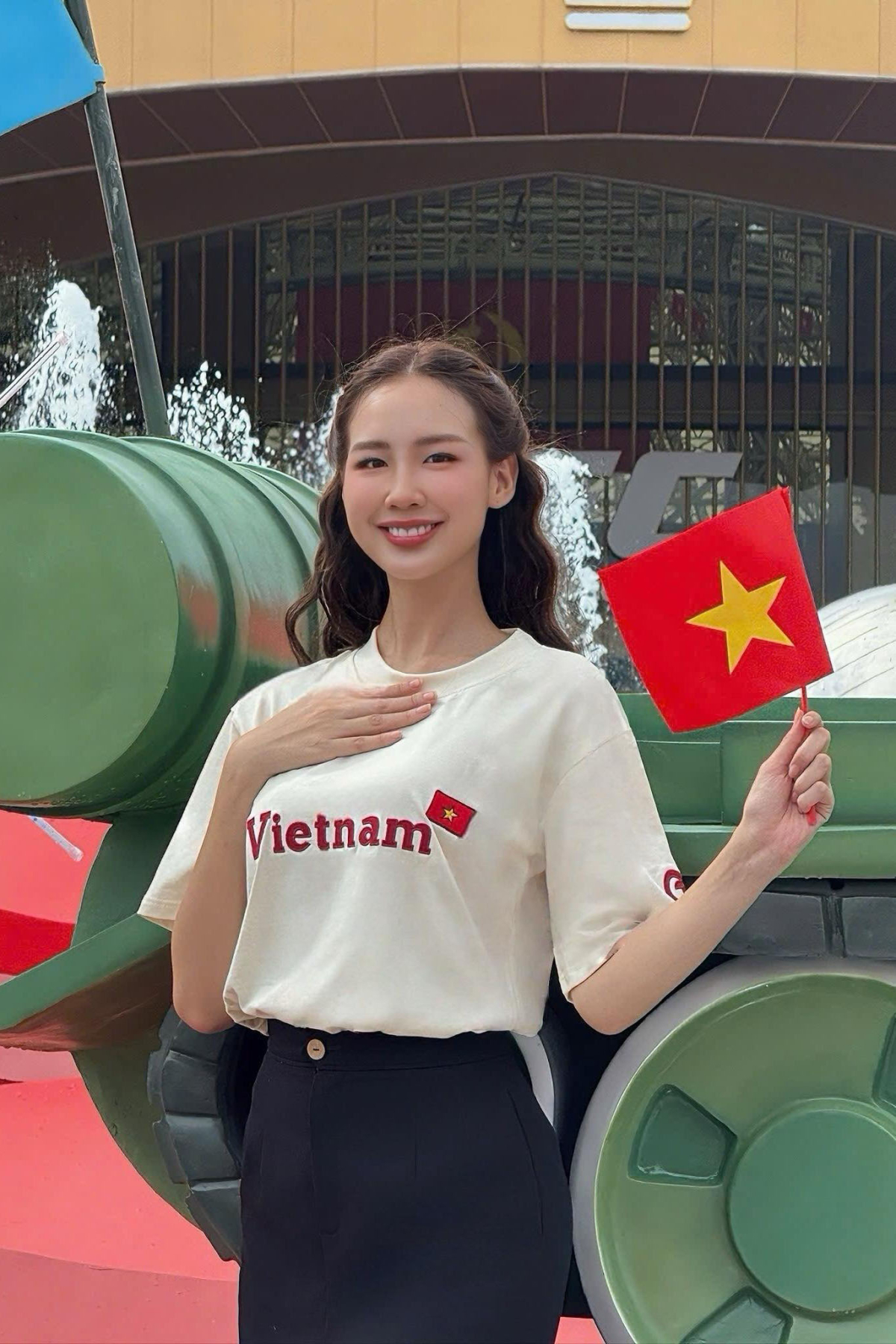 Hoa hậu Bảo Ngọc thể hiện một phong cách trẻ trung, hiện đại nhưng vẫn đậm tinh thần dân tộc. Cô diện áo thun trắng với dòng chữ Vietnam nổi bật cùng lá cờ đỏ sao vàng – biểu tượng tự hào của đất nước. Chiếc áo đơn giản nhưng mang tính biểu trưng cao, được kết hợp hài hòa với chân váy cạp cao màu đen, tạo nên sự thanh lịch và gọn gàng.  Tóc được uốn nhẹ, buông xõa tự nhiên, cùng nụ cười tươi tắn làm tăng thêm vẻ dịu dàng và gần gũi. Lá cờ nhỏ cầm tay là điểm nhấn sinh động, góp phần hoàn thiện hình ảnh một người phụ nữ trẻ trung, yêu nước và tràn đầy năng lượng tích cực.