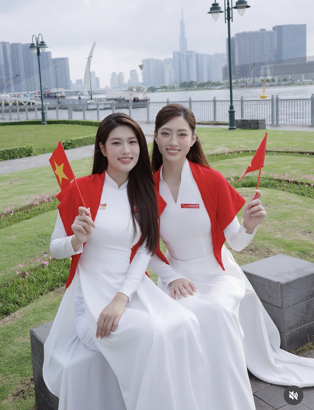 hai nàng hậu Đoàn Thiên Ân và Lương Thùy Linh cùng khoác lên mình tà áo dài trắng tinh khôi – biểu tượng của vẻ đẹp truyền thống, dịu dàng và nền nã của người phụ nữ Việt Nam. Chiếc khăn đỏ nổi bật được choàng khéo léo trên vai, cùng với lá cờ Tổ quốc trên tay, thể hiện tinh thần tự hào dân tộc tràn đầy trong dịp Tết Độc Lập.
