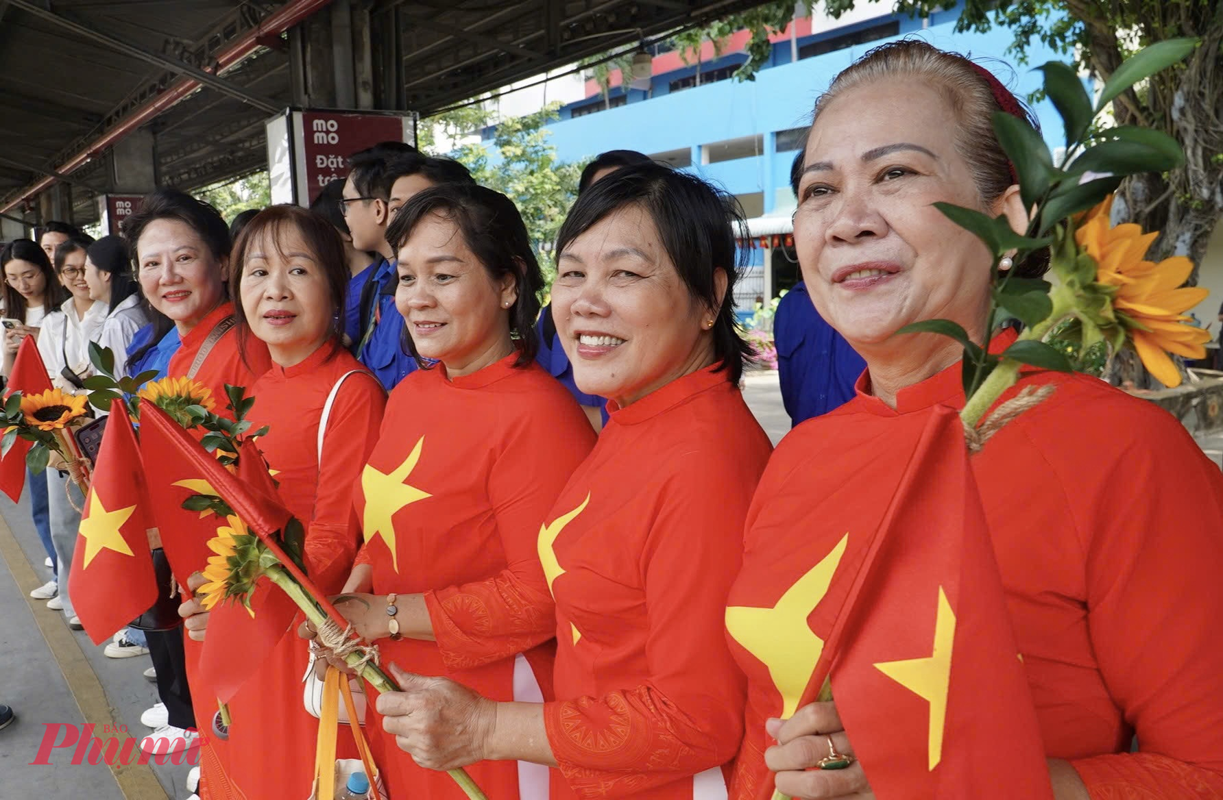 Người dân khoác áo dài đỏ sao vàng, tay cầm hoa và cờ Tổ quốc, ánh mắt rạng ngời chờ đợi giây phút gặp mặt - Ảnh: Thanh Tâm
