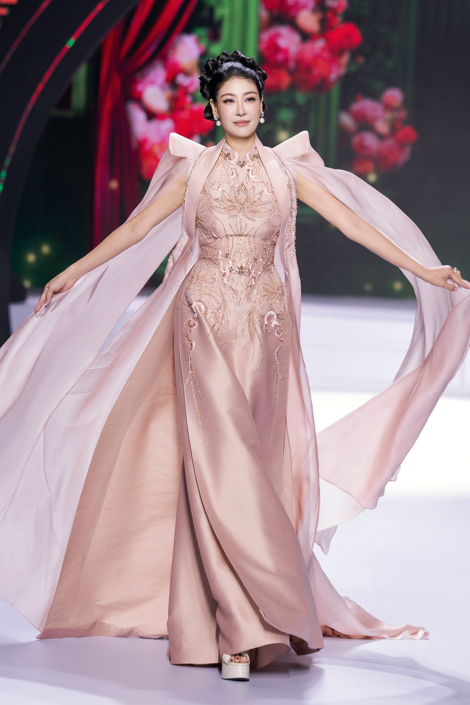 Hà Kiều Anh xuất hiện đầy thanh lịch và kiêu sa trên sàn catwalk với thiết kế áo dài cách tân màu hồng phấn dịu nhẹ. Bộ trang phục mang phong cách hiện đại nhưng vẫn giữ được nét truyền thống, nổi bật với họa tiết hoa thêu tinh xảo kết hợp đính kết lấp lánh, tạo điểm nhấn sang trọng và quý phái. Phần vai áo được thiết kế cách điệu với cầu vai cao và tà áo bay bổng như dải lụa mềm, mang lại cảm giác nhẹ nhàng, thanh thoát trong từng bước di chuyển.