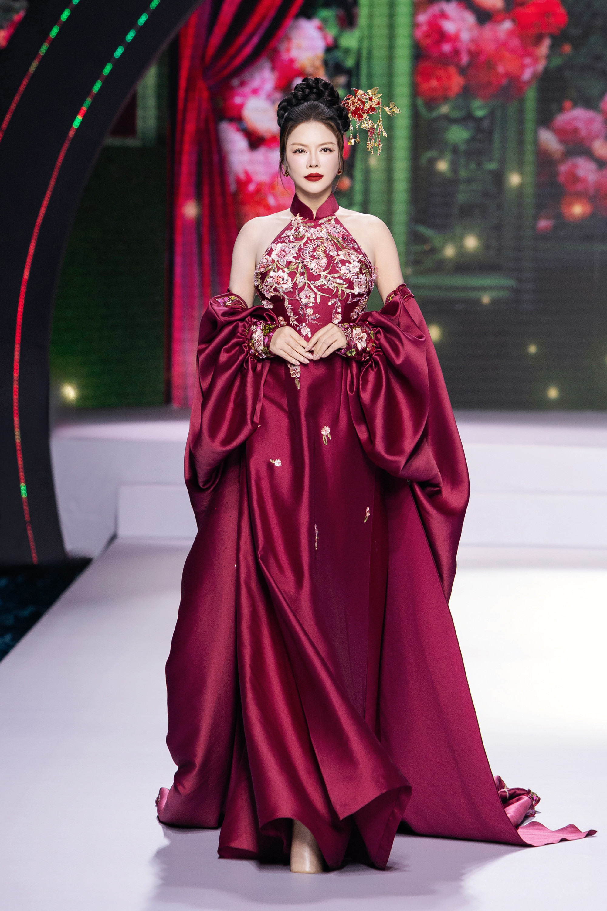 Lý Nhã Kỳ xuất hiện đầy quyền lực và kiêu sa trên sàn catwalk với thiết kế dạ hội màu đỏ rượu vang. Bộ váy mang phom dáng cổ yếm, phần ngực được đính kết cầu kỳ với hoa văn thêu nổi và họa tiết ánh kim, tạo nên vẻ đẹp vừa cổ điển vừa hiện đại. Điểm nhấn ấn tượng nằm ở tay áo phồng dáng cape, được xử lý bằng chất liệu lụa bóng sang trọng, làm nổi bật sự lộng lẫy của tổng thể trang phục.