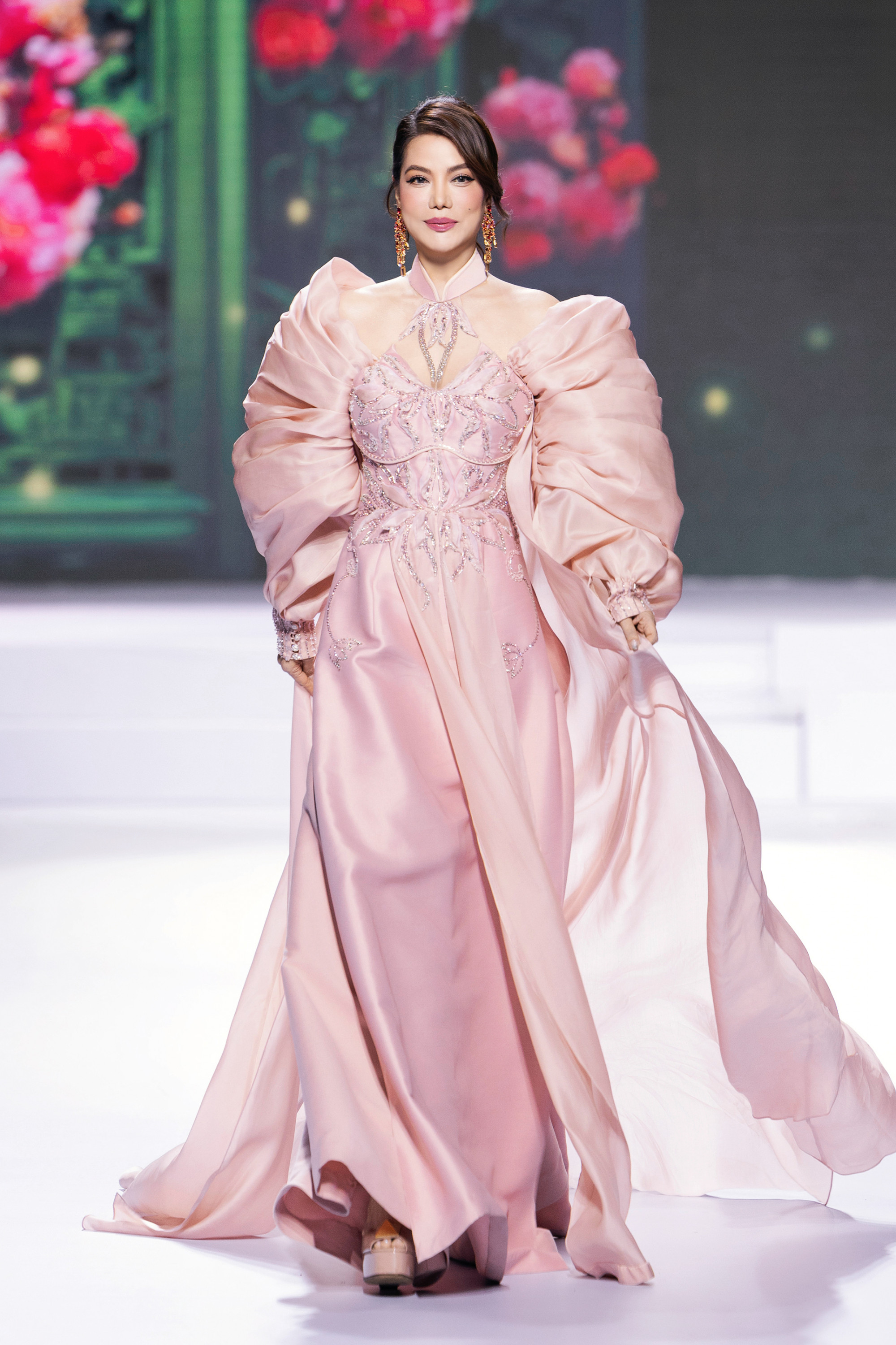 Trương Ngọc Ánh thể hiện phong cách thời trang đầy quyến rũ và quyền lực khi sải bước trên sàn catwalk. Cô diện một bộ váy dạ hội màu hồng pastel thanh nhã, được thiết kế cầu kỳ với phần thân áo đính đá lấp lánh và các họa tiết thêu tinh xảo, tôn lên vẻ đẹp sang trọng và nữ tính. Điểm nhấn ấn tượng nhất là phần tay áo phồng lớn mang hơi hướng cổ điển, tạo nên sự độc đáo và thu hút mọi ánh nhìn. Phần cổ yếm cách điệu kết hợp với kiểu tóc búi thấp và hoa tai dài càng làm nổi bật nét kiêu sa và đẳng cấp của nữ diễn viên.