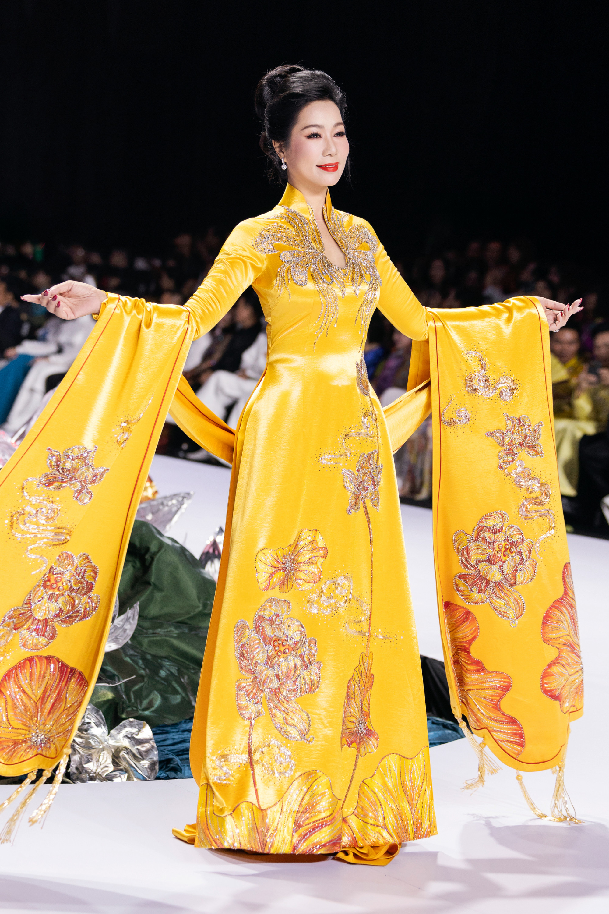 Trịnh Kim Chi toát lên vẻ đẹp quý phái và duyên dáng khi trình diễn trên sàn catwalk với bộ áo dài truyền thống mang phong cách hoàng gia. Thiết kế áo dài màu vàng rực rỡ, chất liệu lụa óng ánh làm tôn lên nét sang trọng và quyền lực. Những họa tiết thêu hoa sen, hoa cúc và các loài hoa truyền thống được đính kết cầu kỳ bằng kim sa lấp lánh, tạo điểm nhấn nổi bật trên thân áo và tà áo.