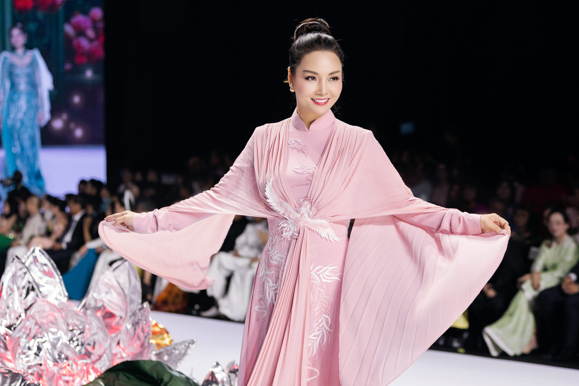 Mai Thu Huyền xuất hiện duyên dáng trong tà áo dài cách tân màu hồng phấn, mang đến vẻ đẹp vừa ngọt ngào vừa sang trọng. Thiết kế nổi bật với chi tiết chim phượng tung cánh được thêu tỉ mỉ trên phần eo, kết hợp những đường xếp ly mềm mại trải dài từ ngực xuống tà áo, tạo hiệu ứng uyển chuyển theo từng bước đi.