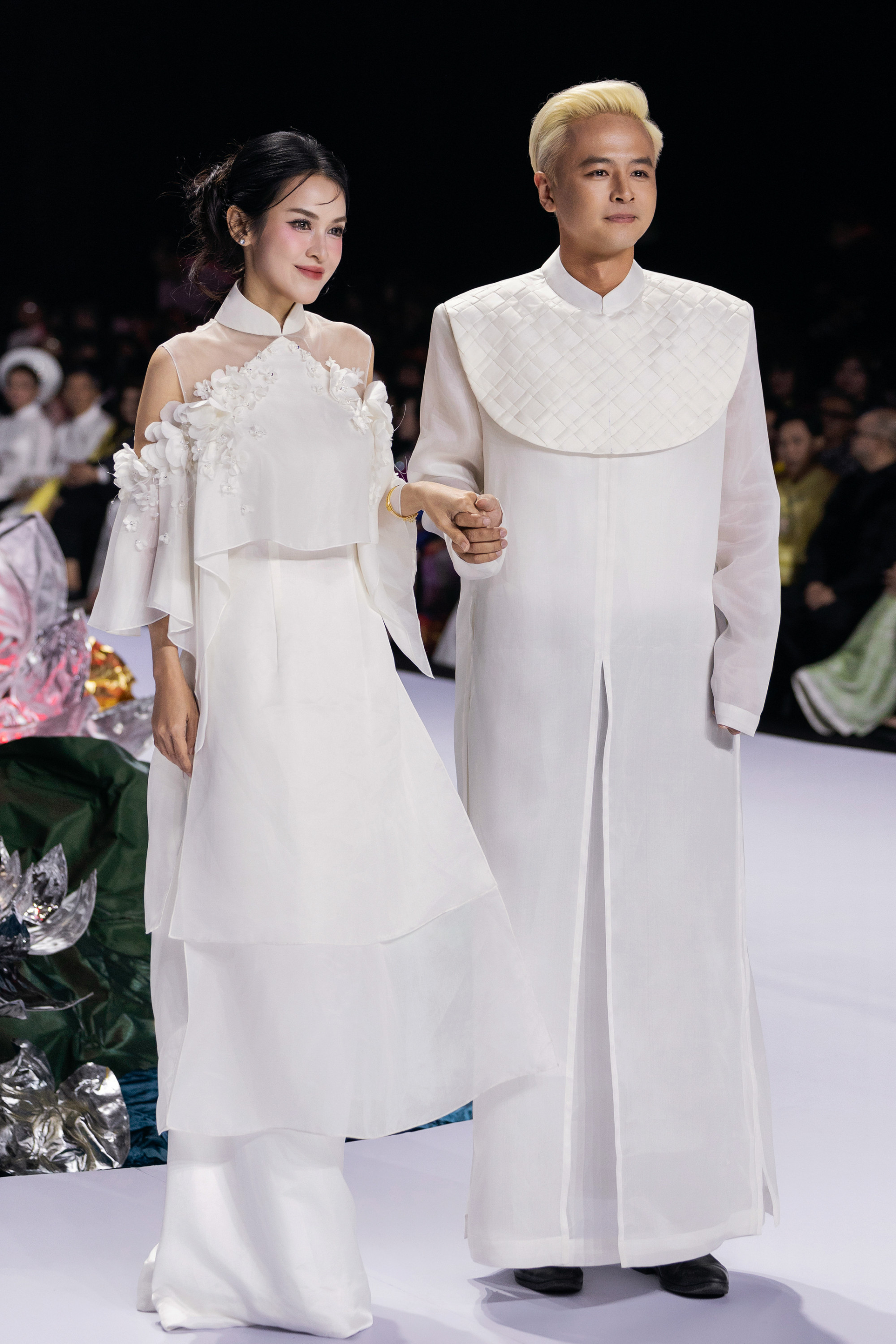 Vợ chồng diễn viên Tú Vi - Văn Anh cùng sánh bước trên sàn catwalk với thiết kế áo dài cưới đồng điệu. Tú Vi diện thiết kế áo dài cách tân với chi tiết vai lệch và lớp voan trong suốt, điểm nhấn là những bông hoa 3D tinh xảo trải dọc phần ngực và tay áo, mang lại cảm giác mềm mại, nữ tính nhưng vẫn hiện đại. Trong khi đó, Văn Anh gây ấn tượng với áo dài nam dáng suông chất liệu mỏng nhẹ, kết hợp phần yếm đắp chéo dạng ô vuông nổi tạo nét phá cách, vừa lạ mắt vừa giữ tinh thần truyền thống.