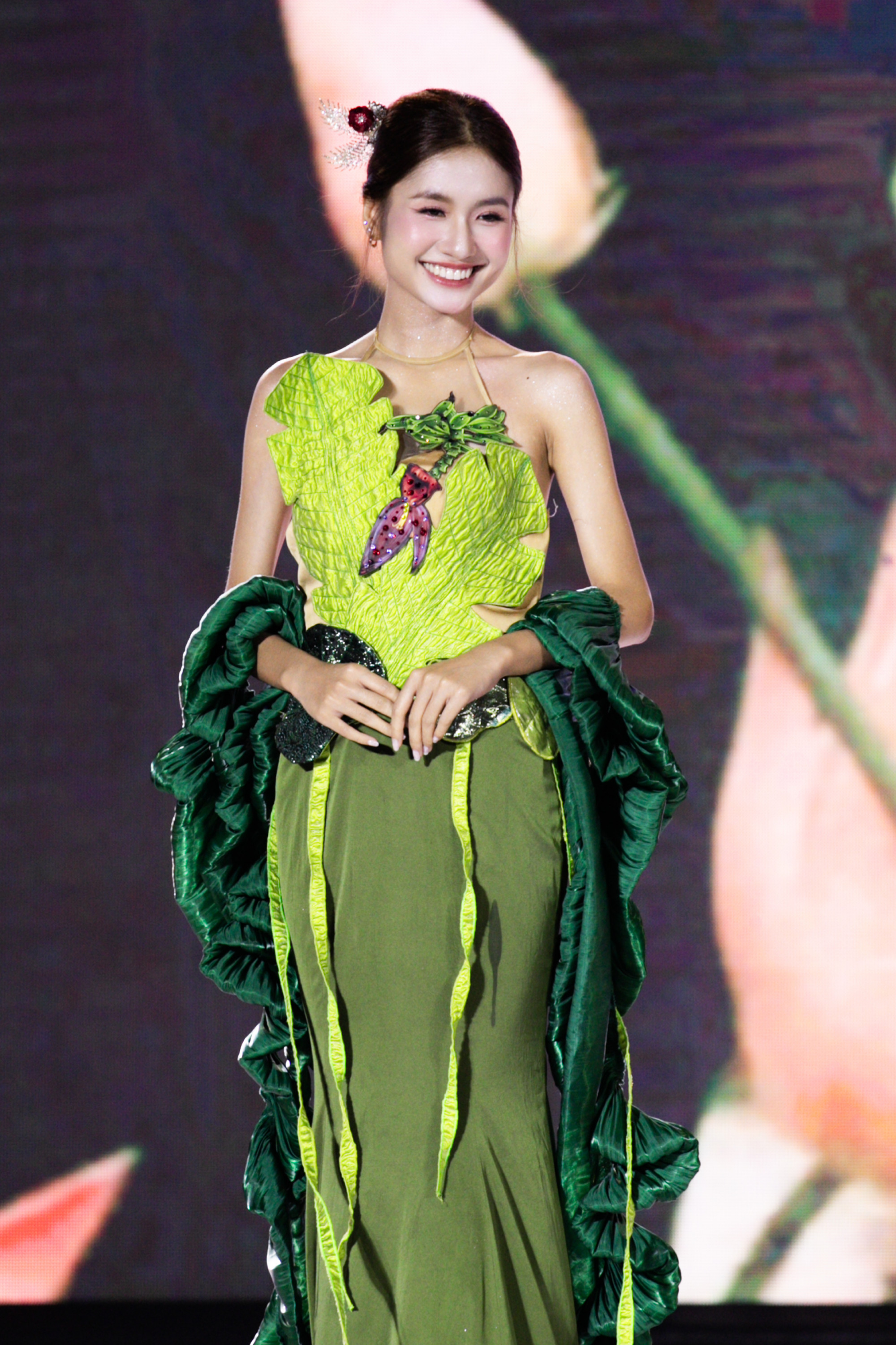 Phần tà váy buông mềm cùng chi tiết tua rủ tạo hiệu ứng chuyển động uyển chuyển, lớp vải xếp nếp ở tay mang lại nét ấn tượng, nghệ thuật. Kiểu tóc búi gọn, điểm nhấn bằng phụ kiện hoa và lông vũ nhỏ giúp tôn lên vẻ tự nhiên, thanh khiết.