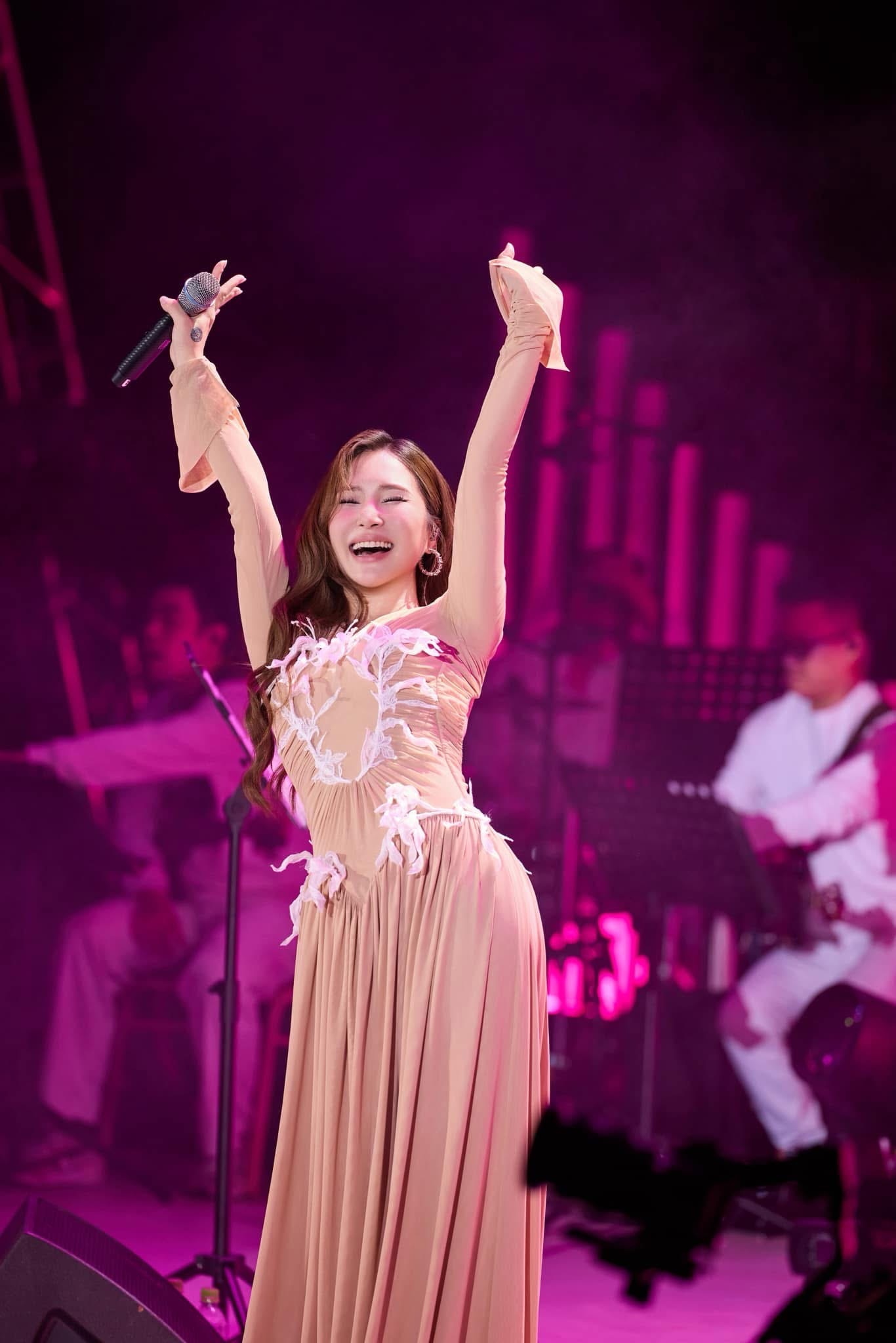 Khoác lên mình nữ ca sĩ chọn chiếc đầm dài màu nude, mang phong cách lãng mạn pha chút hoài cổ. Chất liệu lụa mỏng được xử lý khéo léo, tạo độ rủ nhẹ, ôm lấy từng đường cong tự nhiên. Chi tiết hoa trắng đính nổi trải dọc thân váy như những vệt ký ức mỏng manh, gợi cảm giác dịu dàng nhưng đầy nội lực. Màu nude ấm áp hòa cùng ánh đèn hồng trên sân khấu tạo nên khung cảnh tràn đầy cảm xúc, nơi Hương Tràm như đang hát bằng cả trái tim.