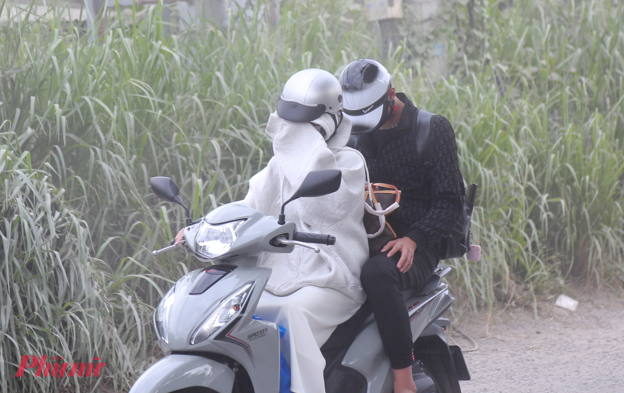 Đường cửa ngõ TPHCM xuống cấp, bụi dày phủ trắng nhà dân như sương mù Cô gái chở bạn phải dùng tay che tạm phía trước để ngăn bụi bay vào mắt.