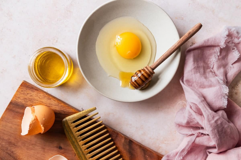 trứng gà là một phương pháp dưỡng phục hồi sâu, đặc biệt phù hợp với mái tóc khô xơ hoặc hư tổn nặng do hóa chất nhuộm, tẩy, uốn. Trứng gà chứa hàm lượng protein, biotin và axit béo tự nhiên dồi dào, giúp tái tạo cấu trúc sợi tóc, tăng độ dày và mang lại vẻ bóng mượt rõ rệt. Khi kết hợp cùng dầu oliu hoặc dầu dừa, hỗn hợp này càng tăng khả năng dưỡng ẩm và làm mềm tóc. Cách thực hiện rất đơn giản: đánh tan 1–2 quả trứng (có thể chỉ dùng lòng đỏ nếu tóc quá khô), trộn với 2–3 thìa dầu dưỡng rồi thoa đều từ chân đến ngọn, massage nhẹ để kích thích lưu thông da đầu. Quấn tóc bằng khăn ấm hoặc mũ trùm và ủ trong 30–40 phút. Cuối cùng, gội sạch bằng nước mát và dầu gội nhẹ để tránh làm chín trứng. Duy trì 1 lần/tuần sẽ giúp mái tóc phục hồi độ đàn hồi, trở nên mềm mượt và chắc khỏe hơn từng ngày.