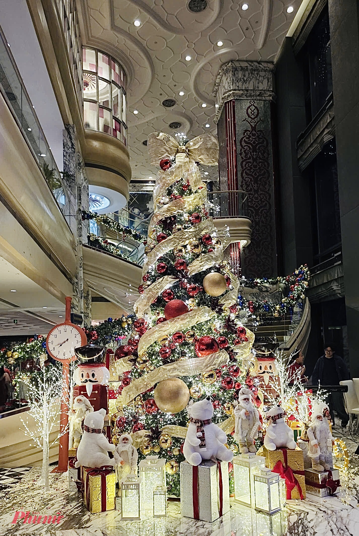 Phong cách trang trí Giáng Sinh tại The Reverie Saigon trong những bức ảnh mang đậm dấu ấn sang trọng, xa hoa và đầy chất nghệ thuật, đúng với tinh thần thượng lưu đặc trưng của khách sạn. Ngay tại sảnh lớn, cây thông khổng lồ được trang hoàng bằng ribbon vàng kim quấn xoắn quanh thân, tạo hiệu ứng chuyển động mềm mại như dải lụa đang bay. Những quả châu đỏ, vàng và ánh kim được sắp đặt dày đặc, phản chiếu ánh sáng lung linh từ hàng trăm bóng đèn LED, khiến cây thông trở thành tâm điểm rực rỡ của toàn bộ không gian.