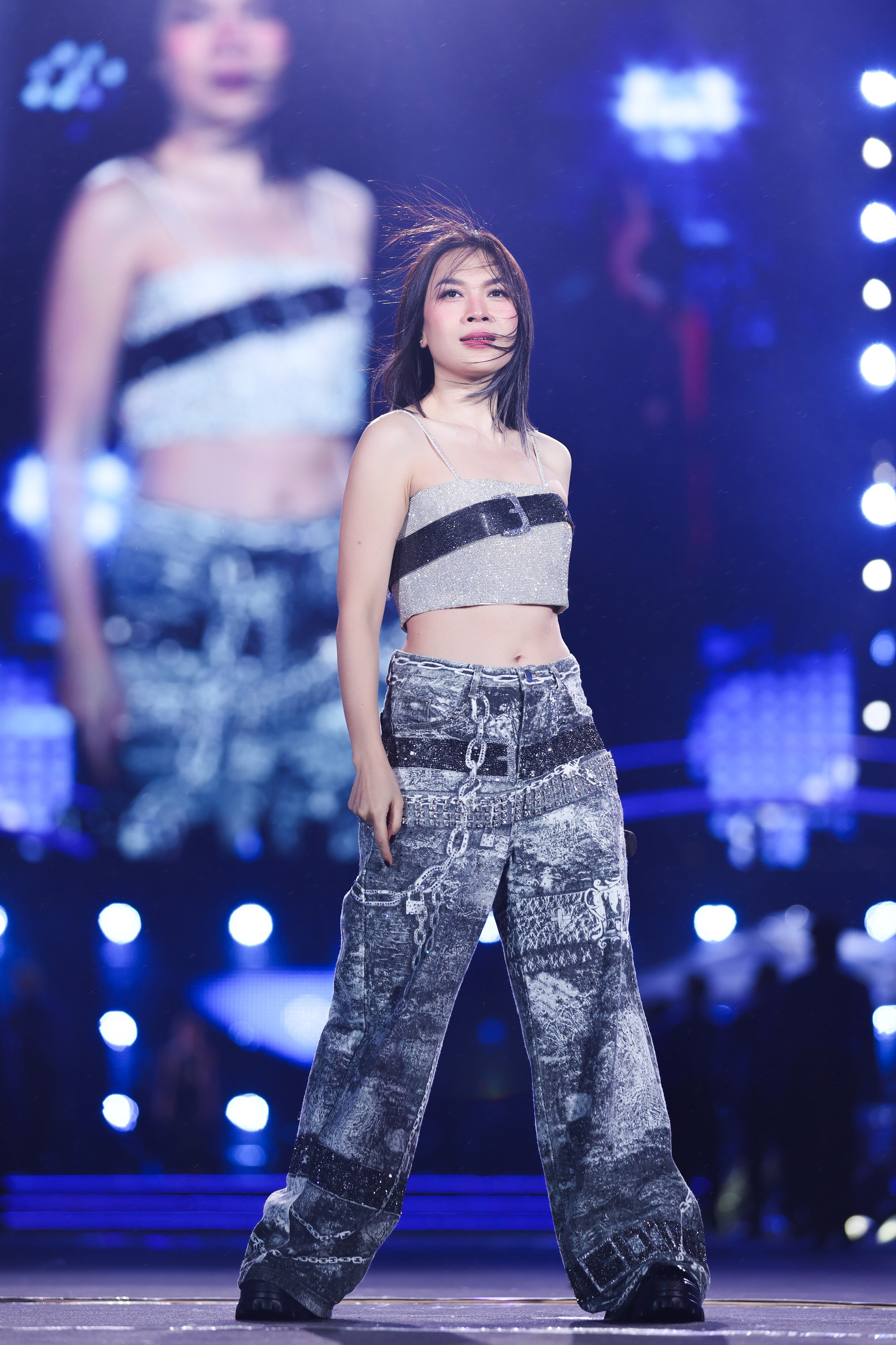 Trang phục croptop ánh bạc phối cùng quần ống rộng họa tiết loang màu tạo cảm giác mạnh mẽ, phóng khoáng nhưng vẫn gợi nét quyến rũ tinh tế cho Mỹ Tâm. Các chi tiết kim loại như dây xích, khóa và dải màu tối giúp tổng thể mang hơi hướng street style pha chất futuristic, rất phù hợp với không khí trình diễn sôi động. Nữ ca sĩ chọn lối trang điểm tự nhiên nhưng sắc nét, lớp nền mỏng, căng bóng giúp da trông khỏe và bắt sáng tốt. Phần mắt được nhấn vừa phải với eyeliner gọn và mi cong, son môi tông hồng nude pha chút ánh bóng mang lại cảm giác trẻ trung.