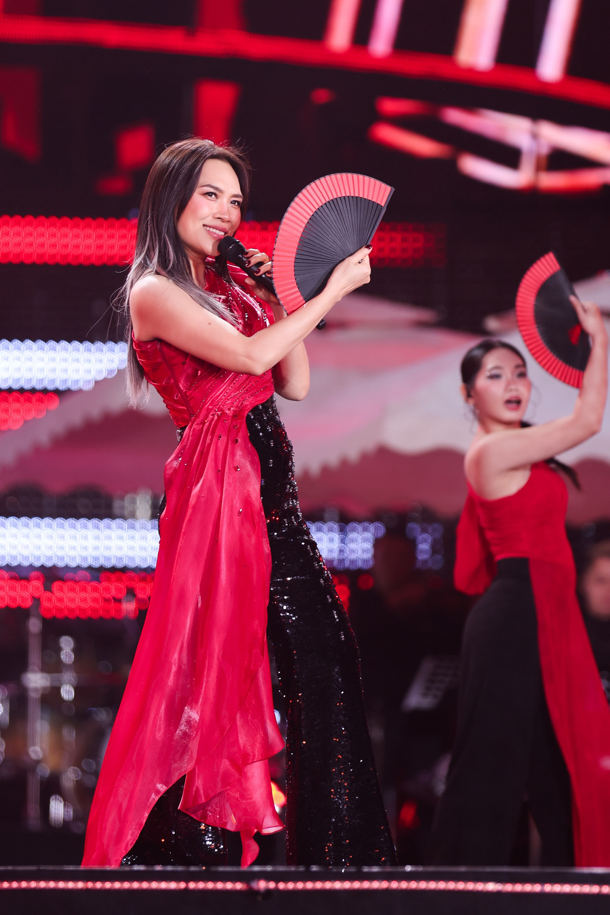 Nữ ca sĩ ghi điểm với phong cách thời trang quyến rũ, mang hơi thở Á Đông hiện đại. Cô lựa chọn bảng màu đỏ - đen tương phản mạnh, tạo hiệu ứng thị giác ấn tượng dưới ánh đèn sân khấu. Thiết kế váy ôm sát với phần thân đen đính sequin lấp lánh giúp tôn dáng và tăng vẻ sang trọng, trong khi lớp vải đỏ mềm mại phủ lệch một bên mang lại cảm giác uyển chuyển, bay bổng theo từng chuyển động. Kiểu dáng không quá cầu kỳ nhưng được xử lý tinh tế, phù hợp với những tiết mục sôi động, giàu năng lượng. 
