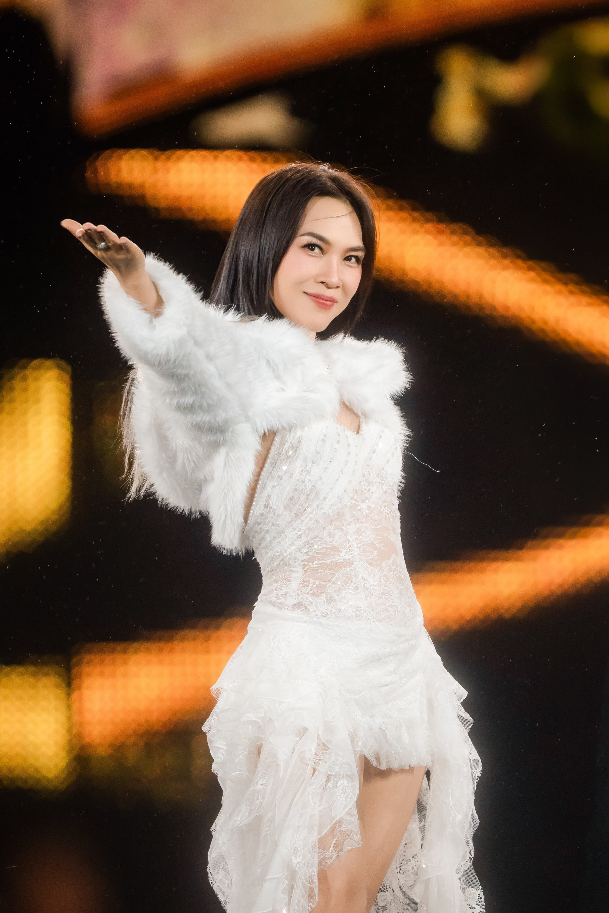 Mỹ Tâm xuất hiện với phong cách thời trang thanh lịch, nữ tính toát lên khí chất ngôi sao. Cô lựa chọn bộ váy trắng ôm sát, chất liệu xuyên thấu nhẹ nhàng kết hợp ren và voan, giúp tôn lên đường nét cơ thể một cách tinh tế mà không phô trương. Phần chân váy được xử lý bất đối xứng, tạo hiệu ứng chuyển động mềm mại, uyển chuyển theo từng cử chỉ. Điểm nhấn nổi bật nằm ở chiếc áo khoác lông trắng khoác hờ trên vai, vừa tăng cảm giác sang trọng, vừa mang hơi hướng cổ điển. Gam màu trắng chủ đạo không chỉ làm nổi bật làn da mà còn góp phần tạo nên tổng thể thuần khiết, trang nhã, phù hợp với không khí trình diễn.