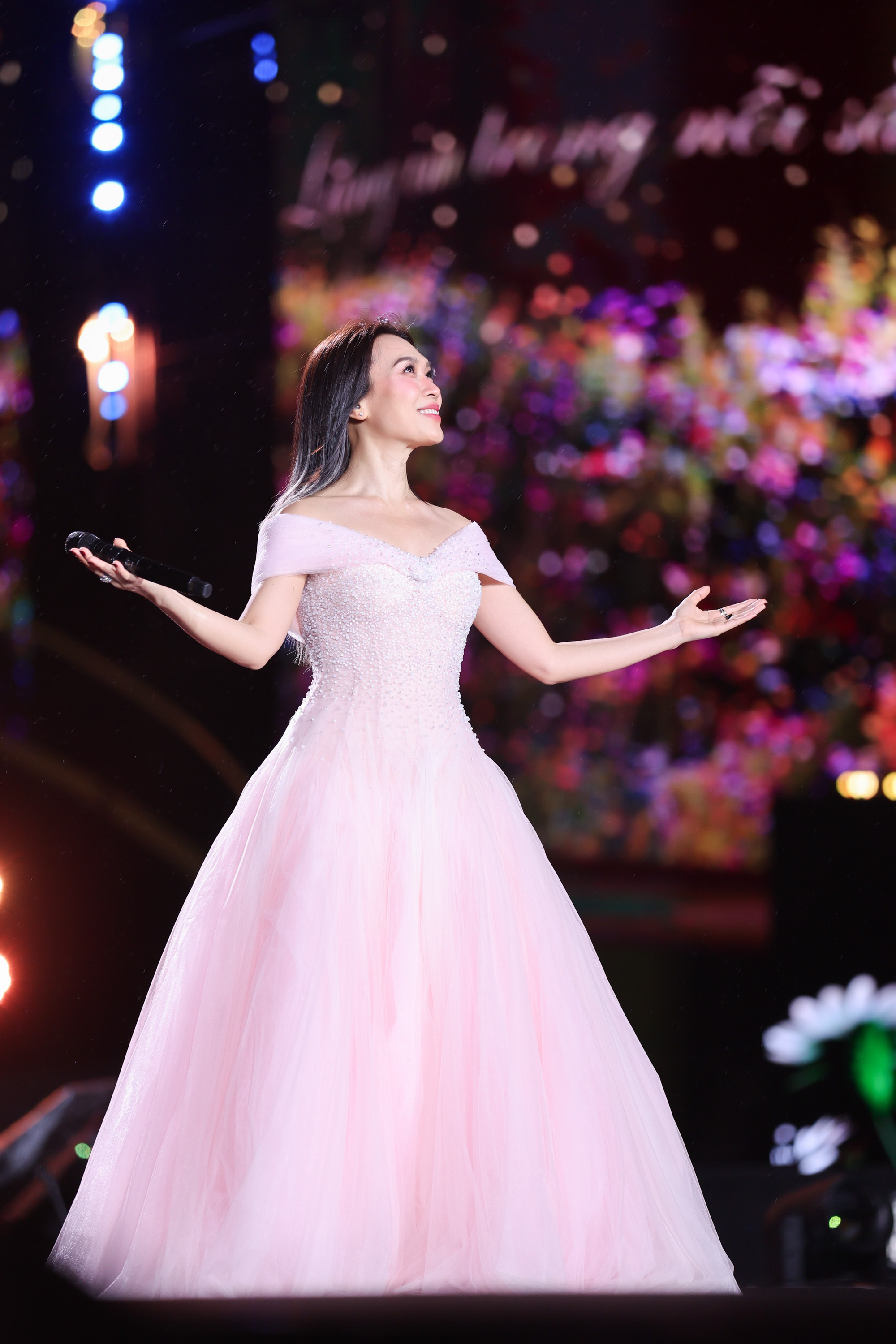 Chiếc váy dạ hội hồng phấn mang phom xòe bồng bềnh, trễ vai tạo cảm giác vừa cổ điển vừa bay bổng. Phần thân trên được đính kết  tinh tế giúp bắt sáng tốt, tôn lên bờ vai và xương quai xanh một cách mềm mại, nữ tính. Chân váy nhiều lớp voan rủ nhẹ, tạo độ chuyển động uyển chuyển theo từng cử chỉ trên sân khấu. Gam hồng pastel không quá rực nhưng nổi bật dưới ánh đèn, mang lại vẻ thanh lịch, dịu dàng và sang trọng, rất phù hợp với những tiết mục mang màu sắc trữ tình, giàu cảm xúc. Mỹ Tâm chọn phong cách trang điểm thiên về lãng mạn, thanh lịch và rất nữ tính, hài hòa với chiếc váy hồng bồng bềnh.
