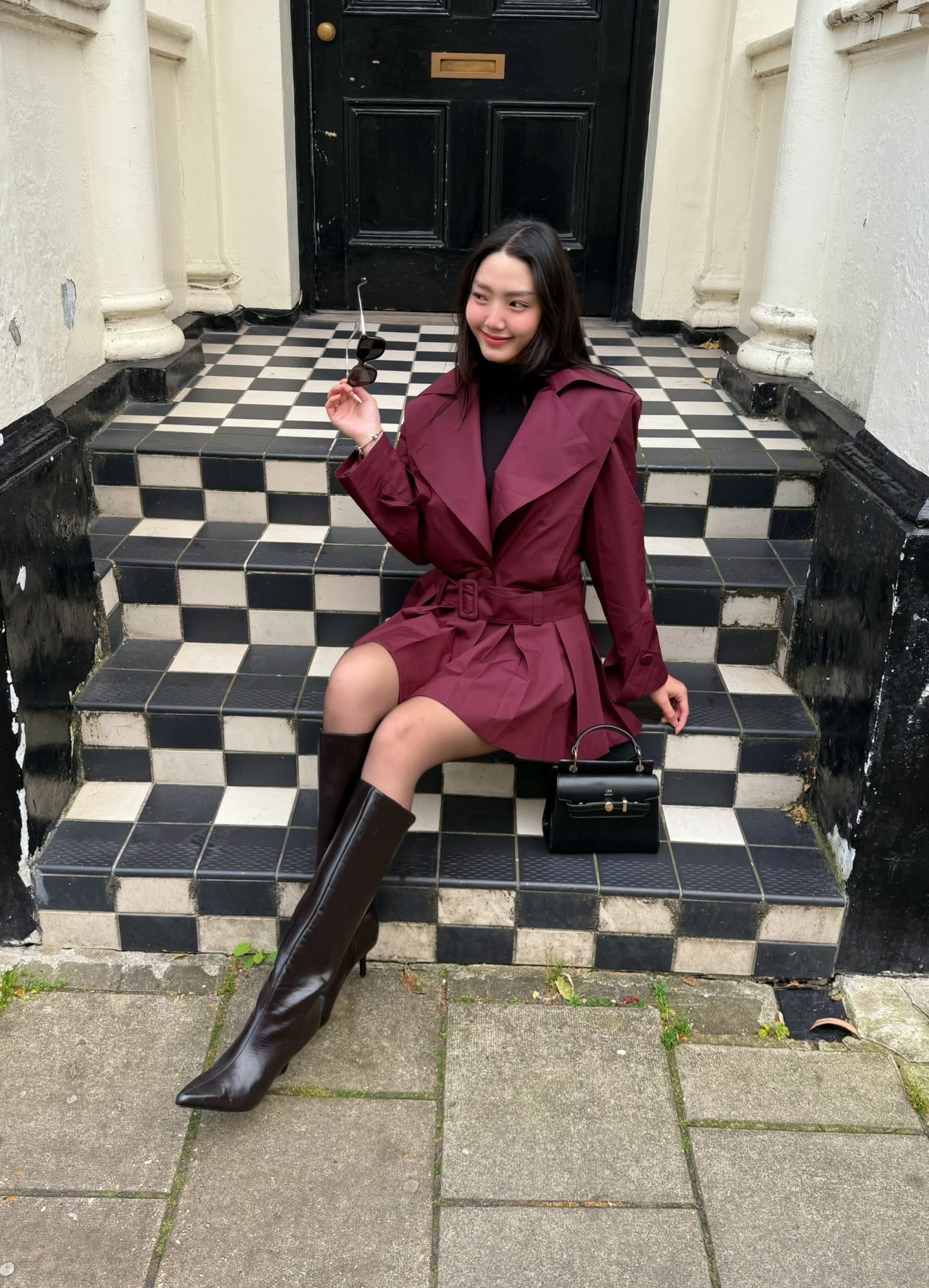 Người đẹp lựa chọn trench coat dáng ngắn màu burgundy, thắt eo gọn gàng, vừa tôn dáng vừa tạo cảm giác sang trọng. Cách phối cùng áo cổ lọ tối màu, boots cao cổ da nâu và túi xách nhỏ cứng cáp mang lại tổng thể chỉn chu, thời thượng. Màu sắc trầm ấm kết hợp bối cảnh kiến trúc cổ điển càng làm nổi bật hình ảnh một cô gái có gu thời trang tinh tế, nữ tính nhưng vẫn đầy bản lĩnh và khí chất.