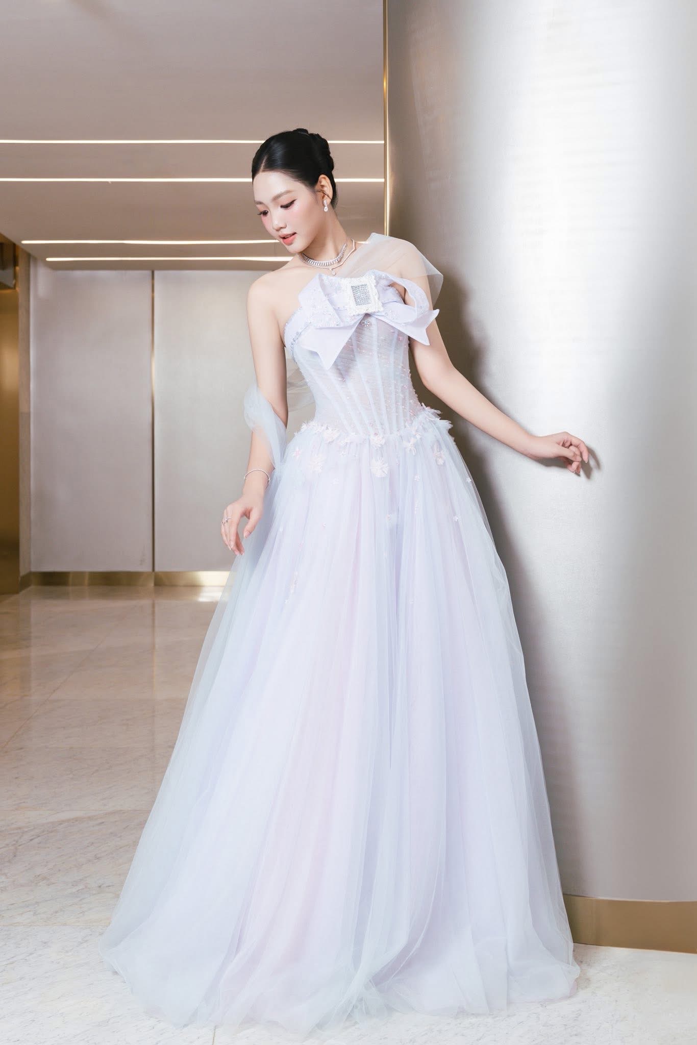 thiết kế váy dạ hội tông trắng pastel với phom dáng corset ôm sát phần thân trên, giúp tôn vòng eo thon gọn và tạo đường nét mềm mại cho cơ thể. Chi tiết nơ lớn đính trước ngực cùng lớp voan mỏng phủ nhẹ vai mang đến cảm giác lãng mạn, tinh khôi, trong khi chân váy xòe dài bằng chất liệu voan nhiều lớp tạo độ bồng bềnh, uyển chuyển theo từng chuyển động.