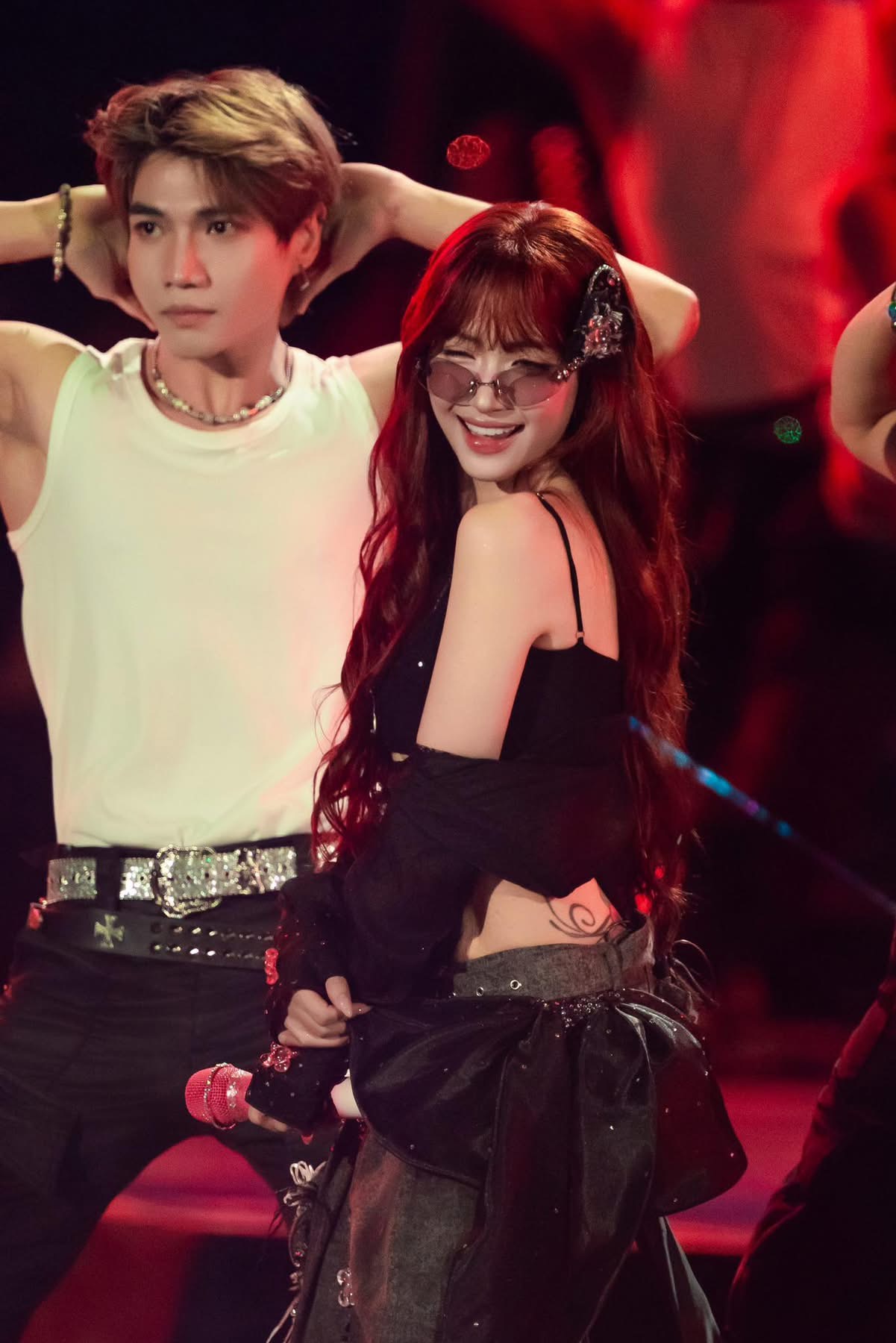 Hòa Minzy diện trang phục tông đen chủ đạo, kết hợp áo 2 dây ôm dáng cùng lớp voan mỏng khoác hờ, tạo cảm giác phóng khoáng và hiện đại. Phần chân váy ngắn phối cùng chi tiết xếp lớp mang hơi hướng street style, giúp tổng thể vừa mạnh mẽ vừa quyến rũ dưới ánh đèn đỏ. Trang điểm được nhấn vào đôi môi đỏ hồng căng mọng và làn da mịn sáng, mắt kẻ sắc nét nhưng không quá đậm, tạo ánh nhìn tự tin, cuốn hút.