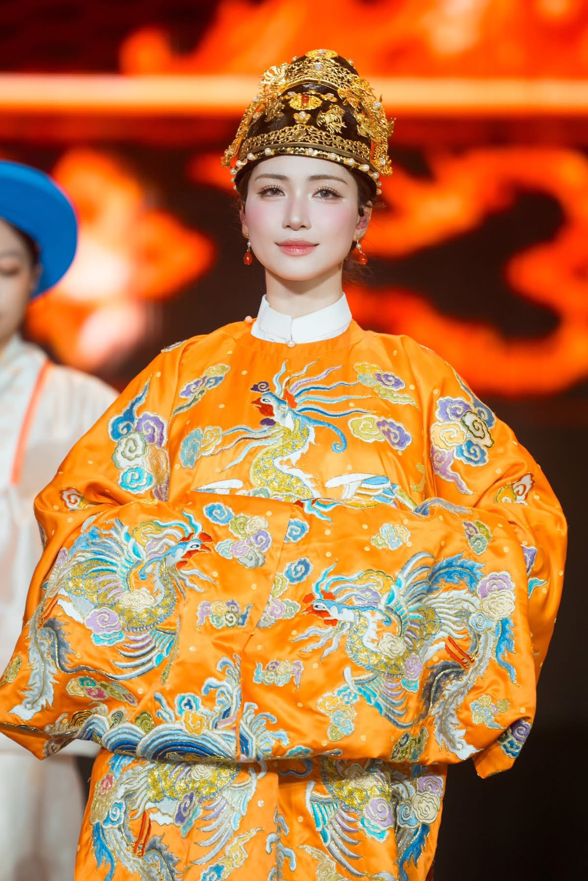 Hòa Minzy gây ấn tượng với phong cách trang phục mang đậm tinh thần truyền thống. Khoác lên mình bộ trang phục gam cam rực rỡ, được thêu họa tiết chim phượng và mây ngũ sắc tinh xảo, gợi liên tưởng đến trang phục cung đình xưa. Phom dáng rộng, tay áo dài tạo cảm giác trang nghiêm, tôn lên vẻ đẹp nền nã. Điểm nhấn đặc biệt là chiếc mấn đội đầu màu đen vàng với chi tiết chạm khắc cầu kỳ, đính đá lấp lánh, góp phần hoàn thiện tổng thể sang trọng. 