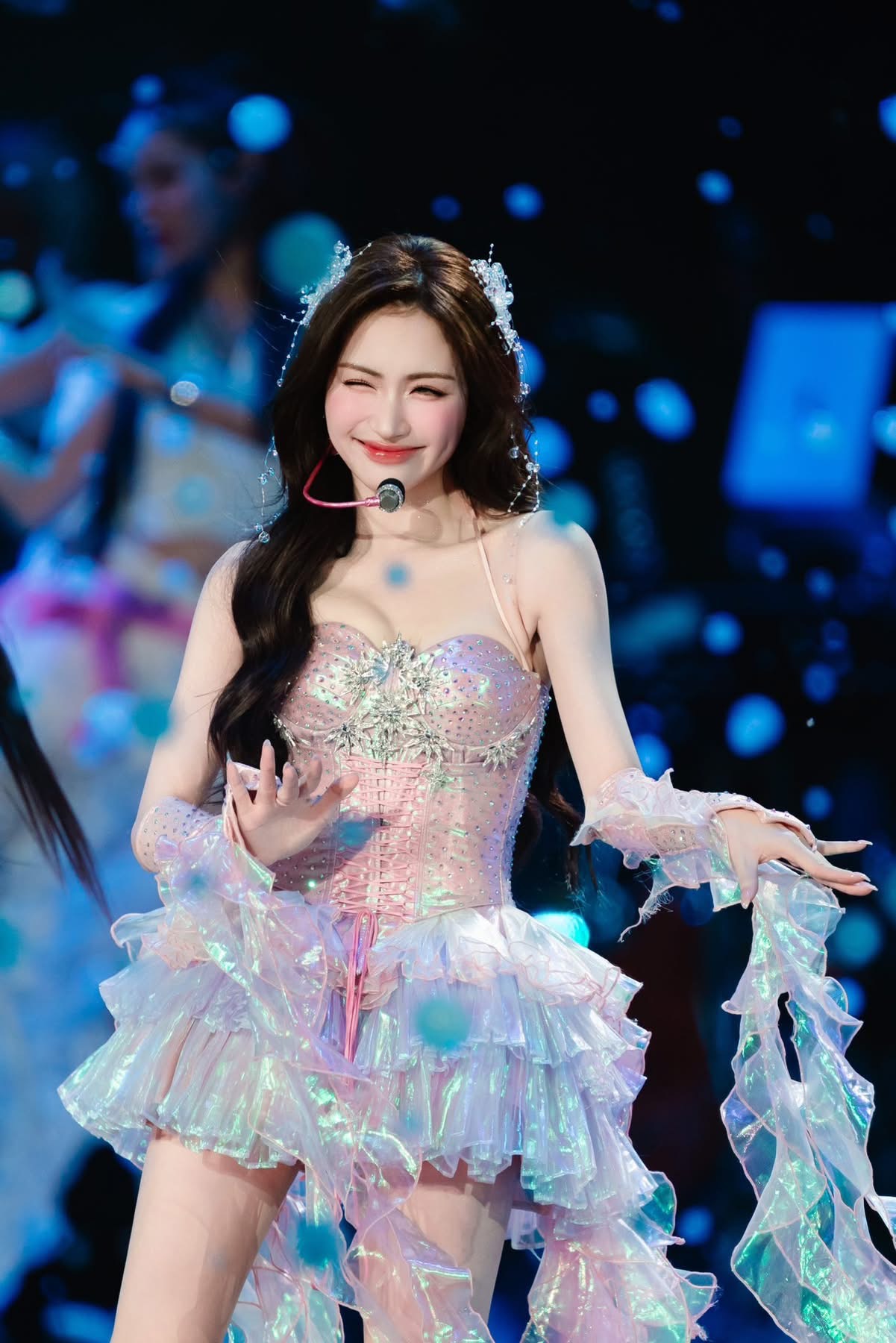 Hòa Minzy mang đến hình ảnh trẻ trung, ngọt ngào pha chút hiện đại sân khấu. Cô diện váy dáng corset màu hồng pastel, chất liệu lấp lánh, kết hợp các tầng bèo nhún mềm mại tạo cảm giác bồng bềnh như công chúa. Thiết kế ôm eo tôn dáng nhưng vẫn giữ nét dễ thương, nữ tính. 