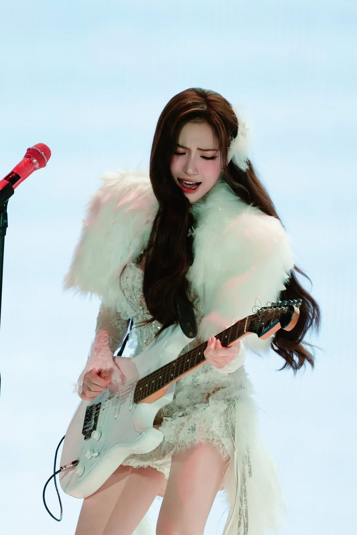 Vơi phong cách sân khấu vừa nữ tính vừa cá tính, Hoà Minzy ghi điểm ấn tượng trong fan concert Bắc Bling. Trang phục tông trắng chủ đạo, kết hợp váy ren xuyên thấu nhẹ cùng áo choàng lông mềm mại, tạo cảm giác sang trọng và bay bổng nhưng vẫn nổi bật khi cầm guitar điện. Thiết kế ngắn, ôm dáng giúp tổng thể trẻ trung, hiện đại hơn. Lối trang điểm ngọt ngào với làn da hồng hào, má ửng nhẹ, môi hồng tươi và đôi mắt được nhấn tinh tế, tôn lên vẻ dịu dàng nhưng vẫn đủ sắc nét dưới ánh đèn sân khấu.