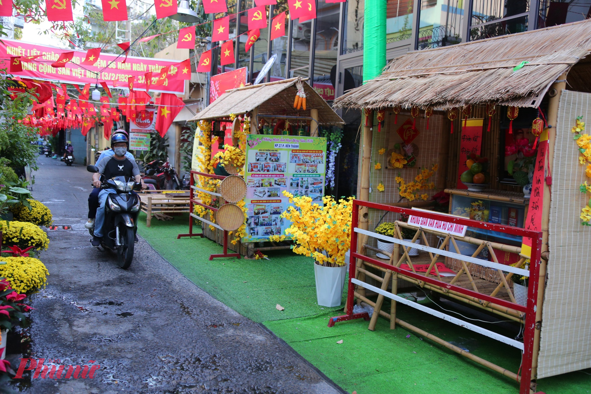 Lối đi được trải thảm cỏ xanh, hai bên là các tiểu cảnh Tết dựng nối tiếp nhau như một “con đường hoa thu nhỏ”. Dọc hai bên hẻm, những chậu cúc vàng được xếp đều tăm tắp, vừa làm đẹp không gian, vừa giữ lối đi gọn gàng, thông thoáng để xe máy vẫn có thể lưu thông