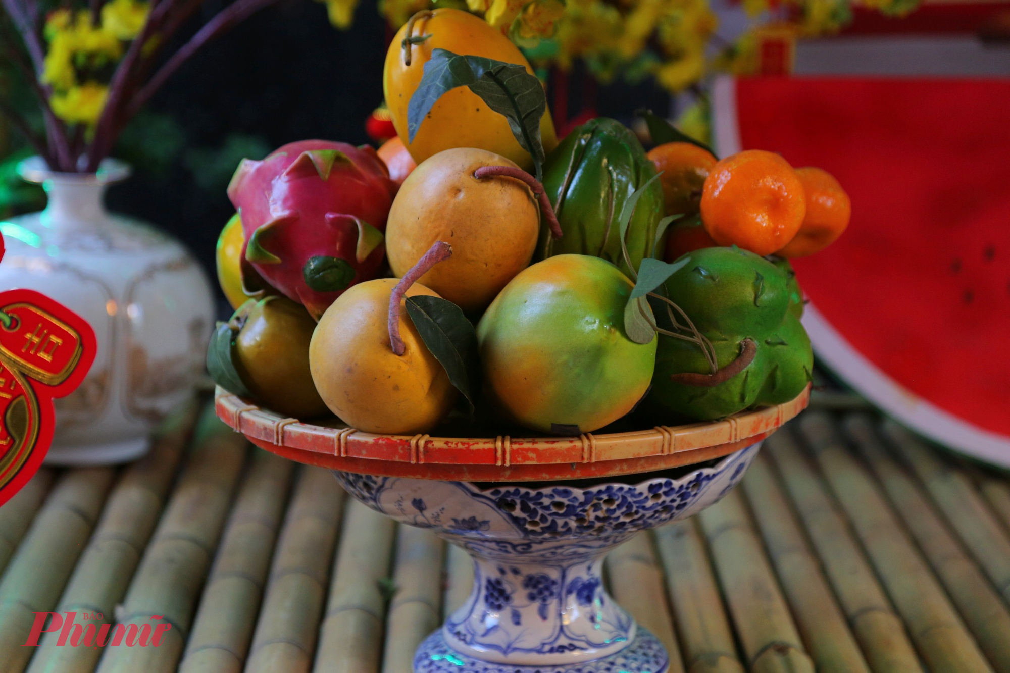 Chính giữa tiểu cảnh là bàn thờ tổ tiên được bày biện gọn gàng với mâm ngũ quả, bình hoa mai vàng, dưa hấu đỏ, bánh mứt… Những vật phẩm quen thuộc này không chỉ mang ý nghĩa cầu mong sung túc, đủ đầy mà còn gợi ký ức tết sum vầy của nhiều thế hệ.