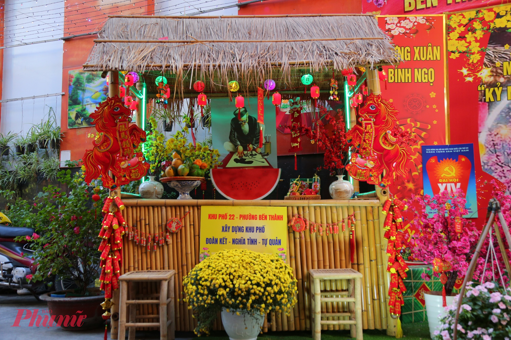 Tiểu cảnh Tết trong hẻm 100 Trần Hưng Đạo được dựng như một gian nhà quê thu nhỏ, đậm chất truyền thống Nam Bộ. Mái lá đơn sơ bằng tranh khô phủ xuống, bên dưới là khung tre mộc mạc, tạo cảm giác thân quen như trở về không gian làng quê ngày tết xưa. Phía sau là phông nền hình ảnh ông đồ ngồi viết thư pháp, tạo điểm nhấn văn hóa, tôn vinh nét đẹp truyền thống mỗi độ xuân về