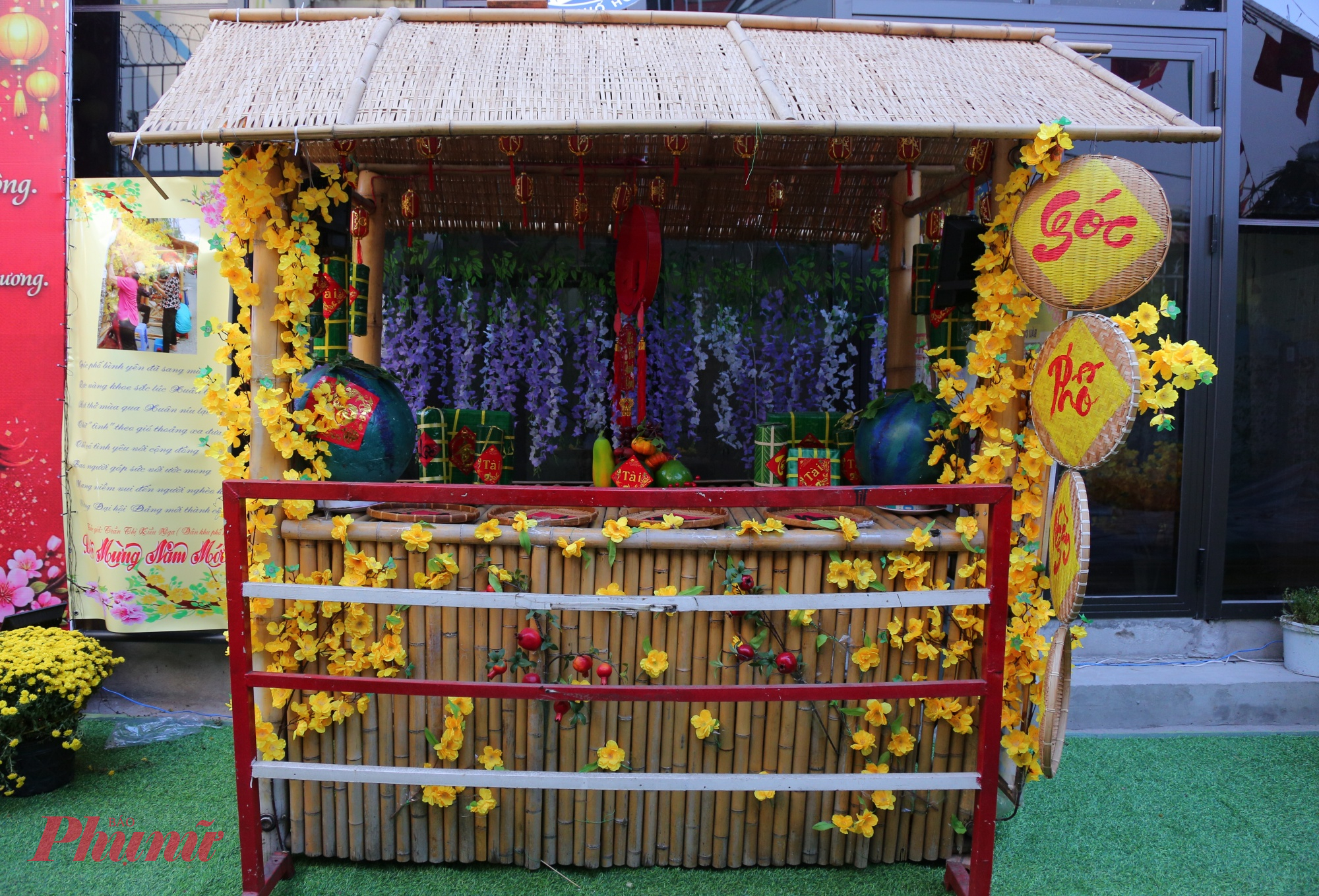 Gian tiểu cảnh được trang trí như “góc chợ Tết - góc quê”, với hàng rào tre, hoa mai vàng kết thành dây leo, những trái dưa hấu xanh, bánh chưng - bánh tét, mâm trái câ. Phía sau là phông nền hoa tím, tạo chiều sâu cho không gian, đồng thời làm nổi bật sắc vàng của hoa mai phía trước. Những chiếc nia tre treo bên hông, bảng chữ “Góc phố tết” mộc mạc gợi cảm giác thân quen, gần gũi như một góc sân nhà ngày tết.