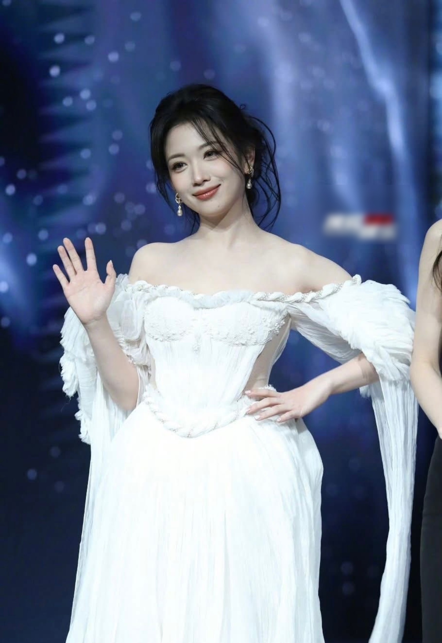 Giữa không gian lấp lánh ánh đèn, Vương Sở Nhiêm xuất hiện trong váy trắng trễ vai, chất liệu voan mềm rủ, tay áo buông dài hờ hững tăng thêm hiệu ứng thị giác. Phom dáng corset ôm nhẹ phần thân trên vừa tôn dáng vừa giữ nét thanh thoát, mang lại cảm giác mong manh nhưng không kém phần sang trọng.