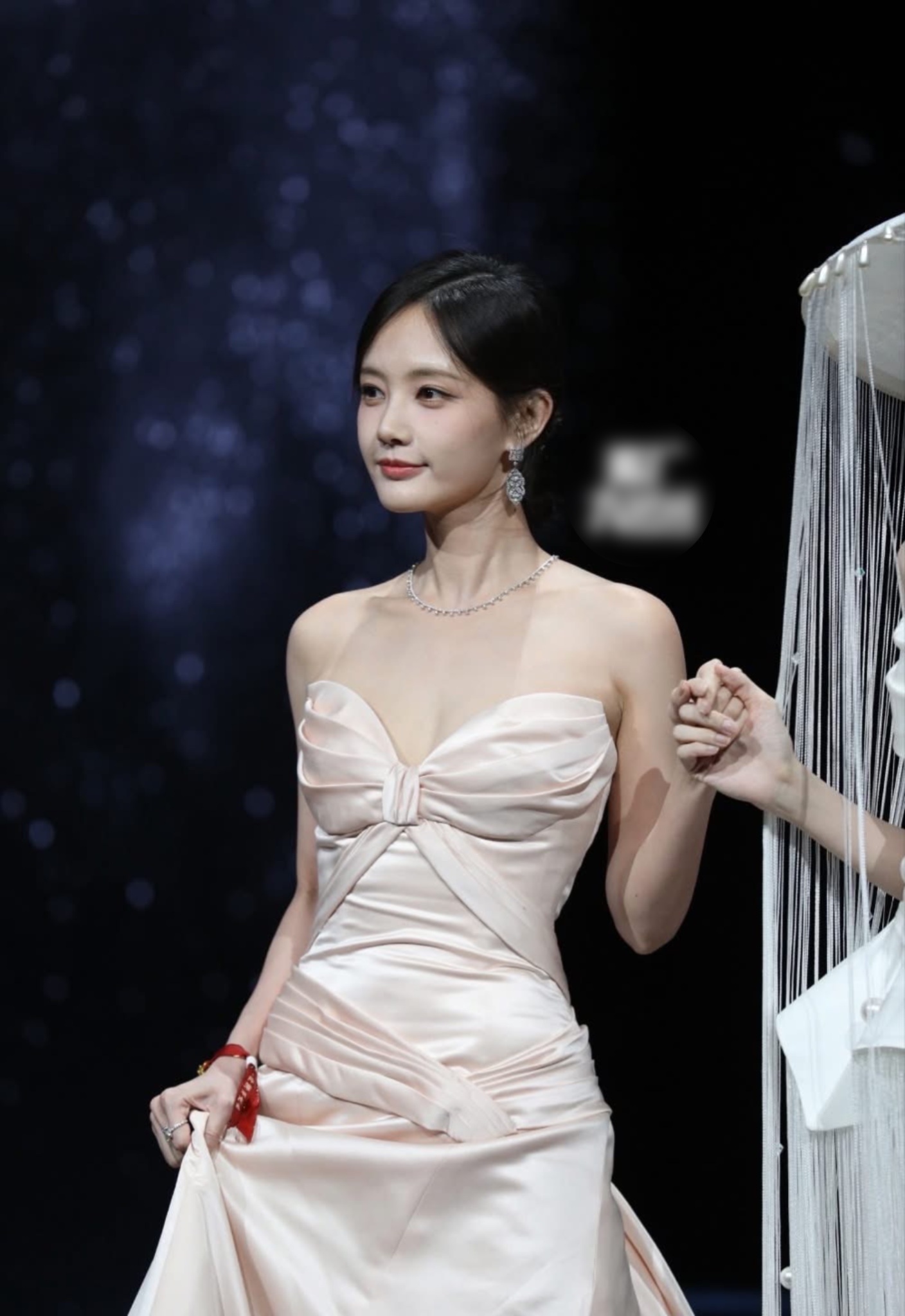 Lý Nhất Đồng ghi điểm với phong cách thảm đỏ thanh lịch pha chút cổ điển, nổi bật nhờ chiếc váy lụa satin màu champagne cúp ngực. Thiết kế ôm sát phần thân trên, nhấn bằng chi tiết xếp nếp mềm mại ở ngực và eo, khéo léo tôn lên vóc dáng mảnh mai và bờ vai trần nữ tính. Cách phối trang sức dây chuyền mảnh và hoa tai dáng rủ cùng kiểu tóc búi thấp gọn gàng càng làm nổi bật khí chất dịu dàng, tinh tế.